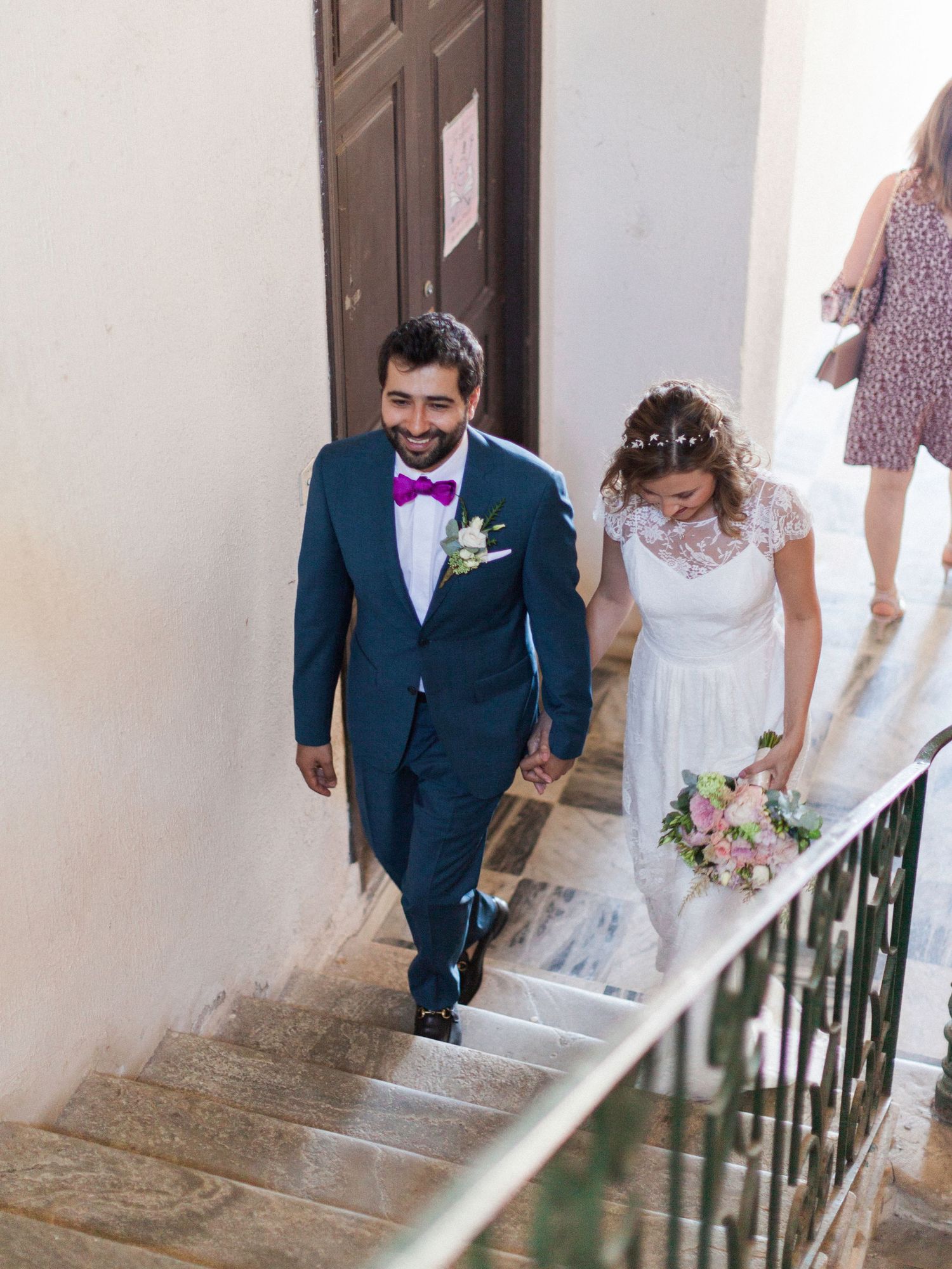 Newlywed couple holding hands walking up stone stairs, groom in blue suit with pink bow tie, bride in white lace dress with bouquet.