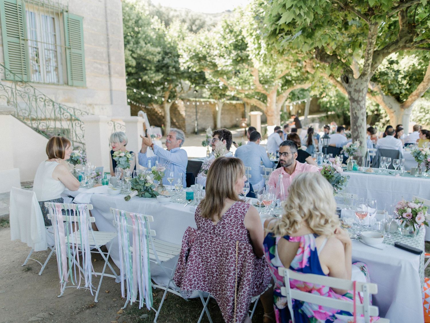 Guests seated at elegant outdoor wedding reception tables in a sunlit Mediterranean garden with white chairs and floral decor.