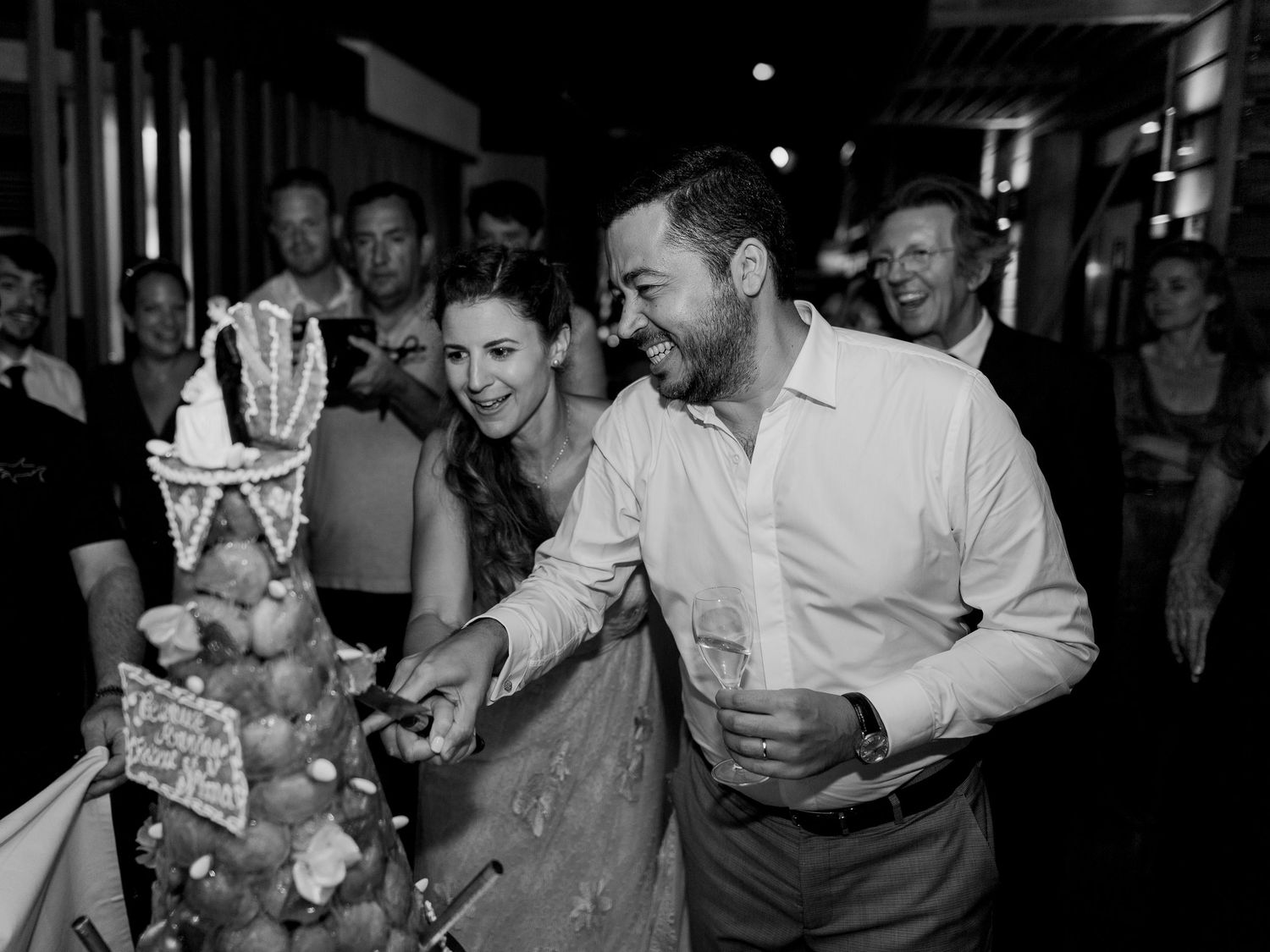 Couple coupant ensemble la pièce montée lors de la réception, invités riant en arrière-plan, photo en noir et blanc au week end ajaccio