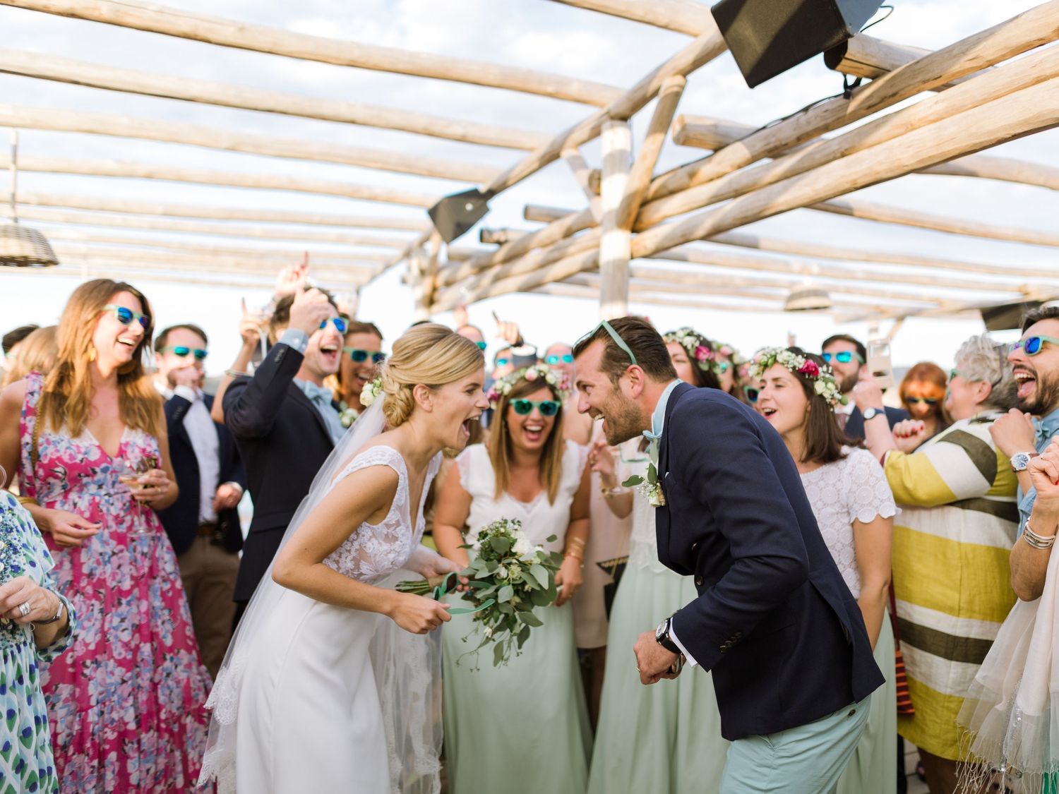 Célébration de mariage joyeuse : les mariés et leurs invités rient sous une pergola rustique en bois lors d'une réception en plein air.