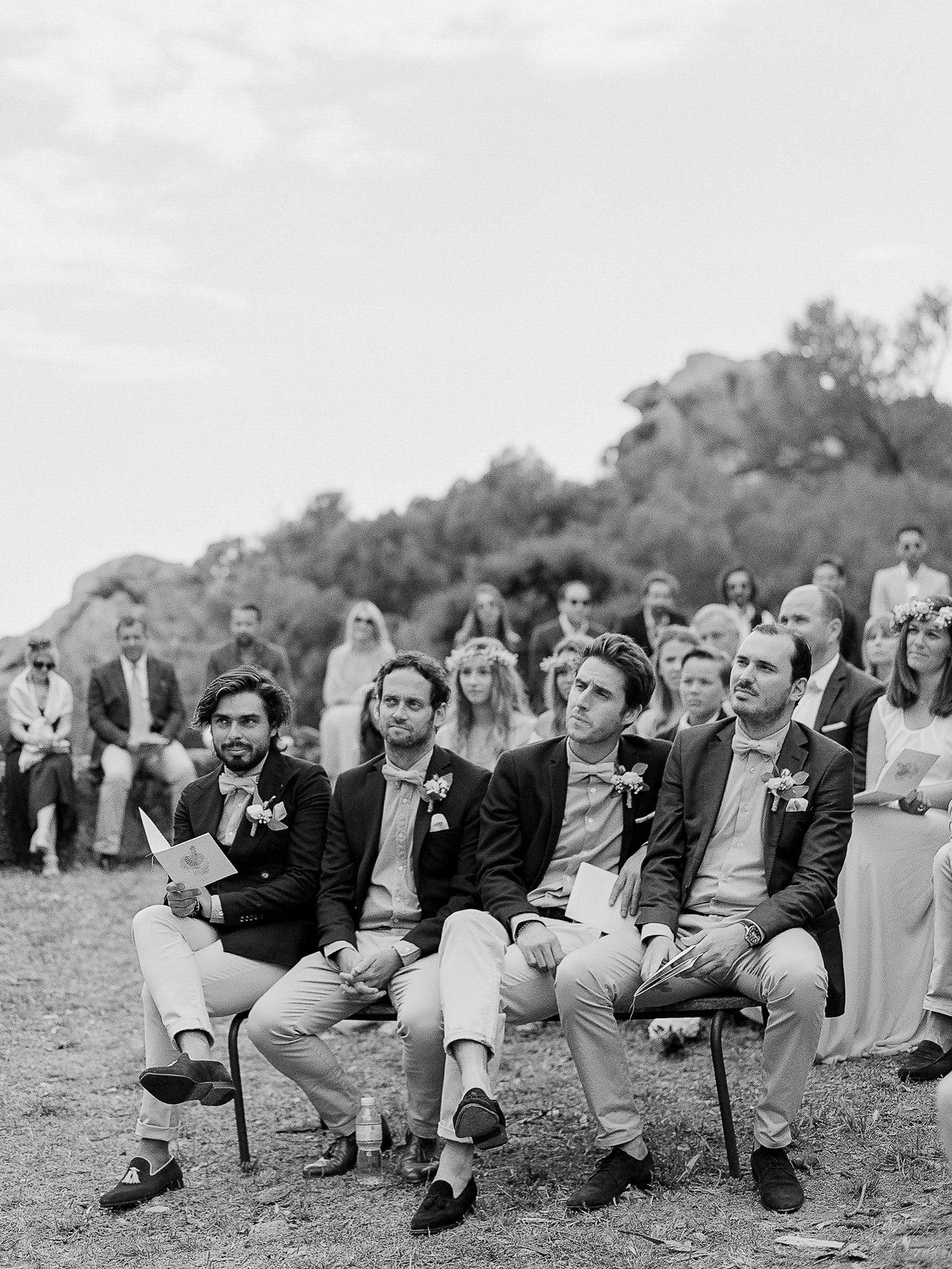 Photo en noir et blanc de garçons d'honneur assis lors d'une cérémonie de mariage en plein air, avec des invités en arrière-plan.