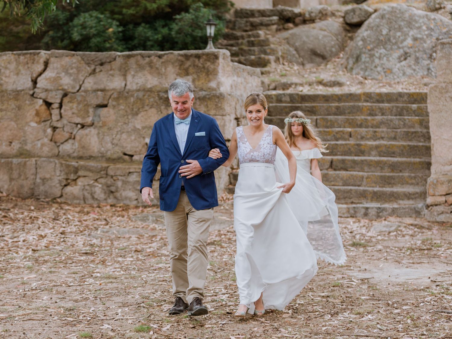 La mariée en robe blanche marche au bras de son père en blazer bleu sur le lieu de la cérémonie, un mariage en plein air avec des ruines de
