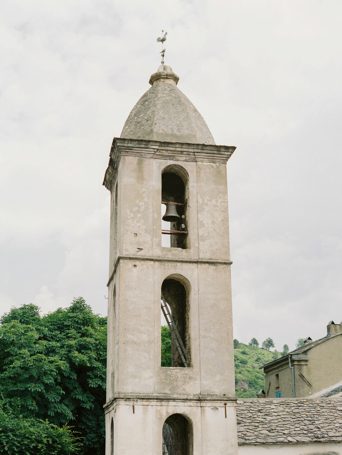 Ancient stone bell tower with arched openings and domed top, surrounded by lush trees in a European village.