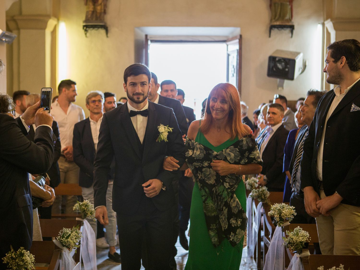 Groom in tuxedo walks down church aisle with woman in green dress during wedding ceremony.