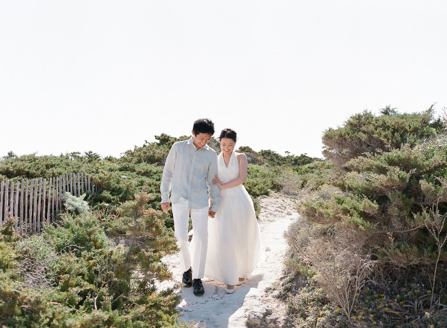 La mariée en robe blanche et le marié en costume bleu clair se promènent main dans la main le long d'un sentier de dunes sablonneuses.