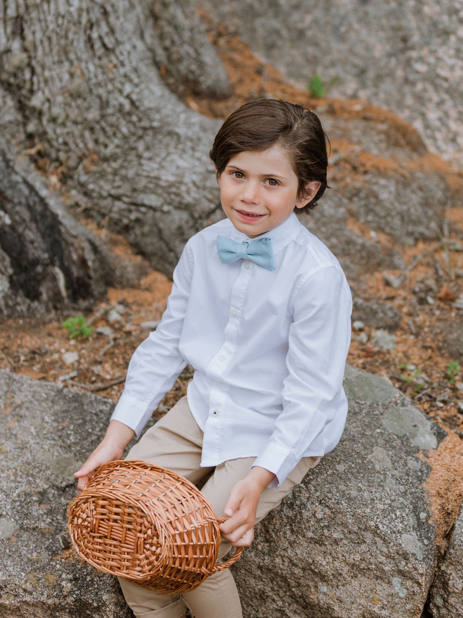 Jeune garçon en chemise blanche et nœud papillon bleu tenant un panier en osier, assis sur des rochers.