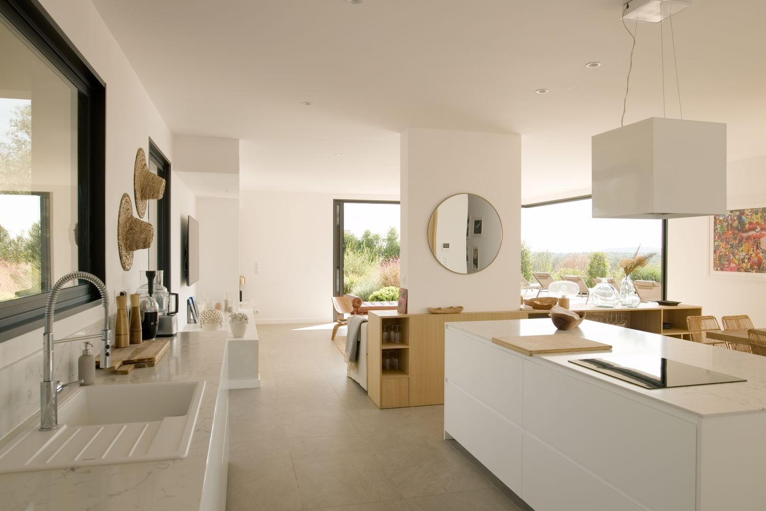 Cuisine moderne blanche ouverte avec îlot central, miroir rond, éléments en bois et vue sur le jardin grâce à de grandes fenêtres.