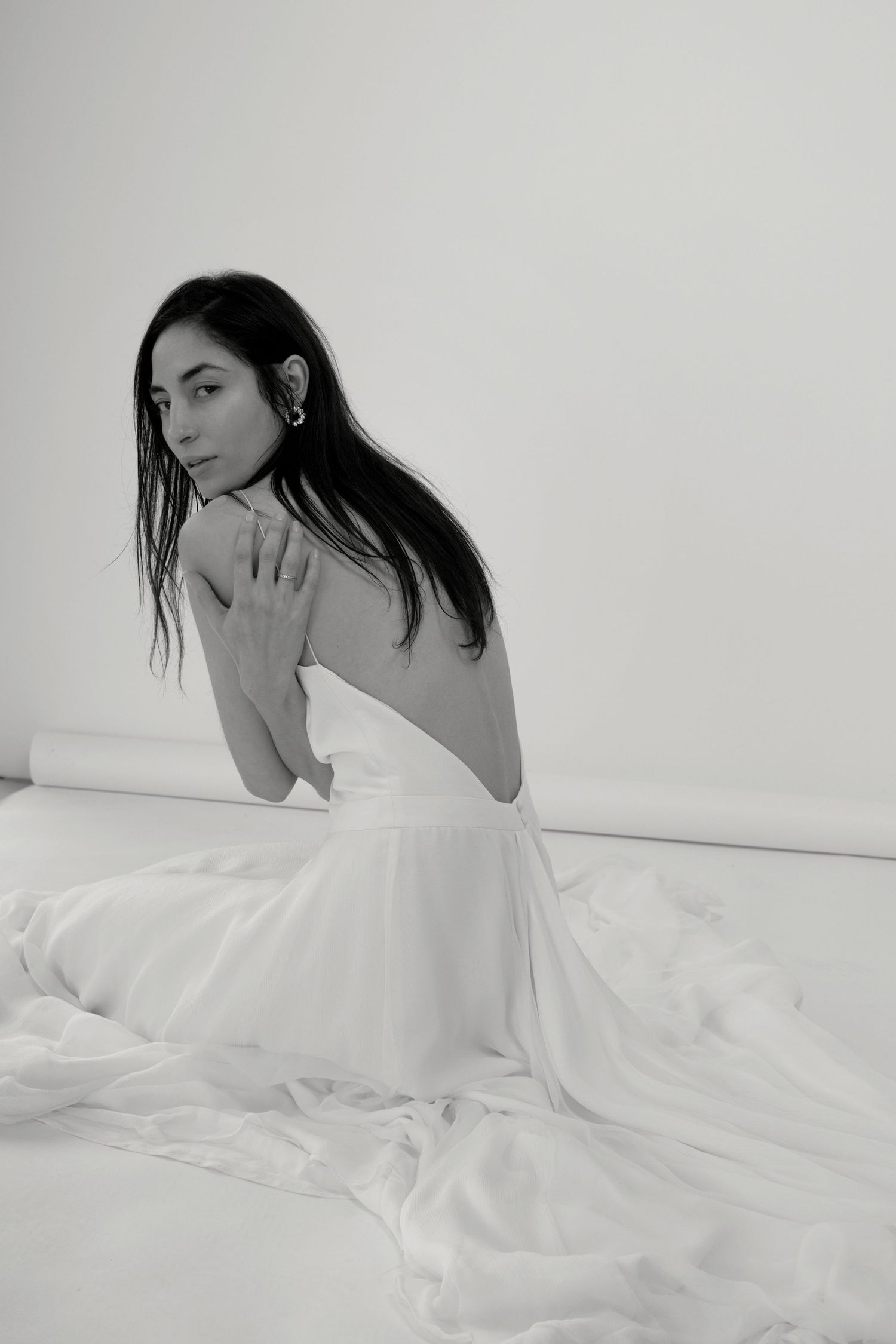 Photographie en noir et blanc d'une femme en robe blanche dos nu, assise avec grâce sur une robe de mariée Caroline takvorian
