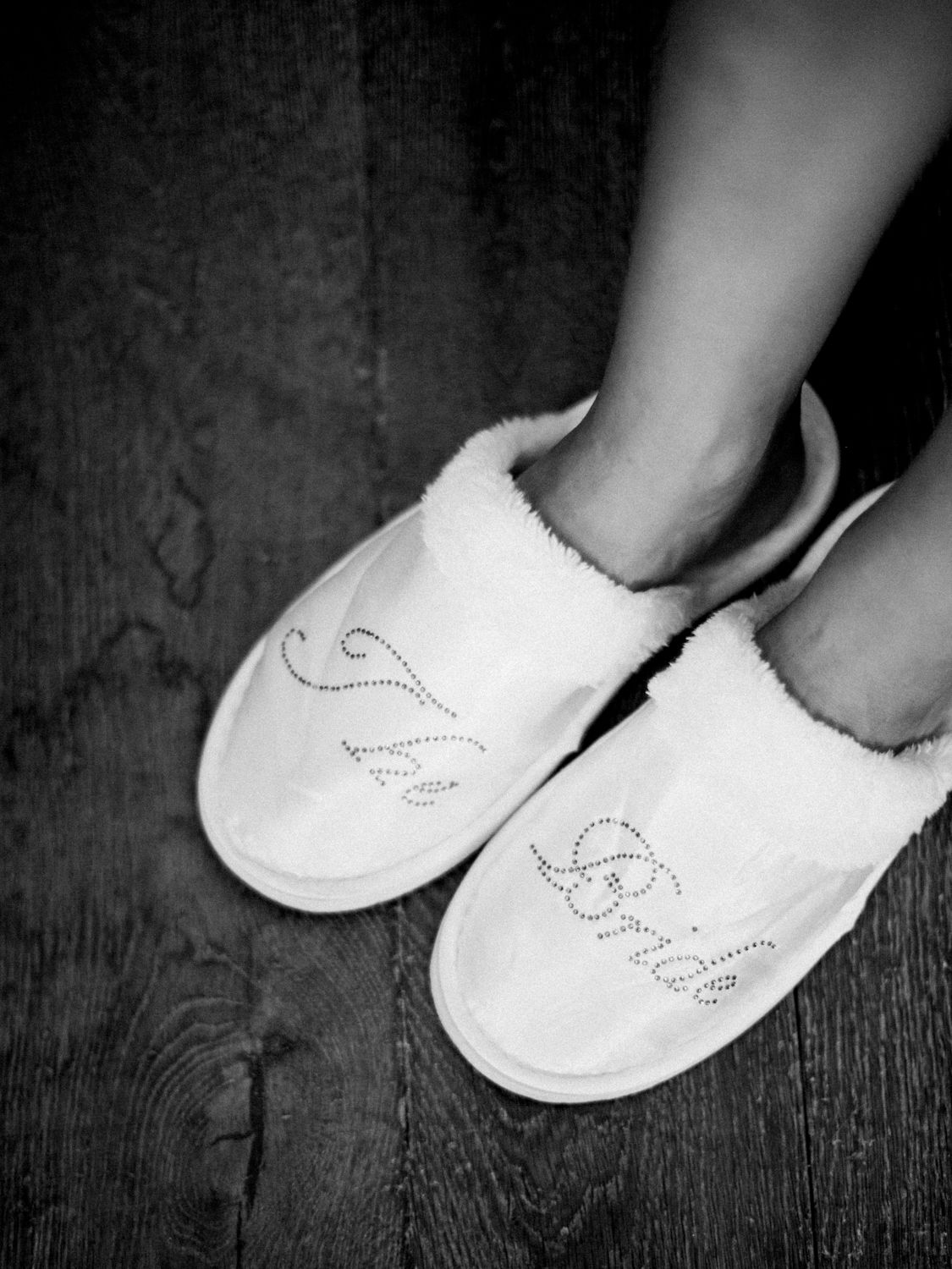 Photo en noir et blanc d'une mariée portant des pantoufles blanches duveteuses à strass sur lesquelles est inscrit « Mariée »