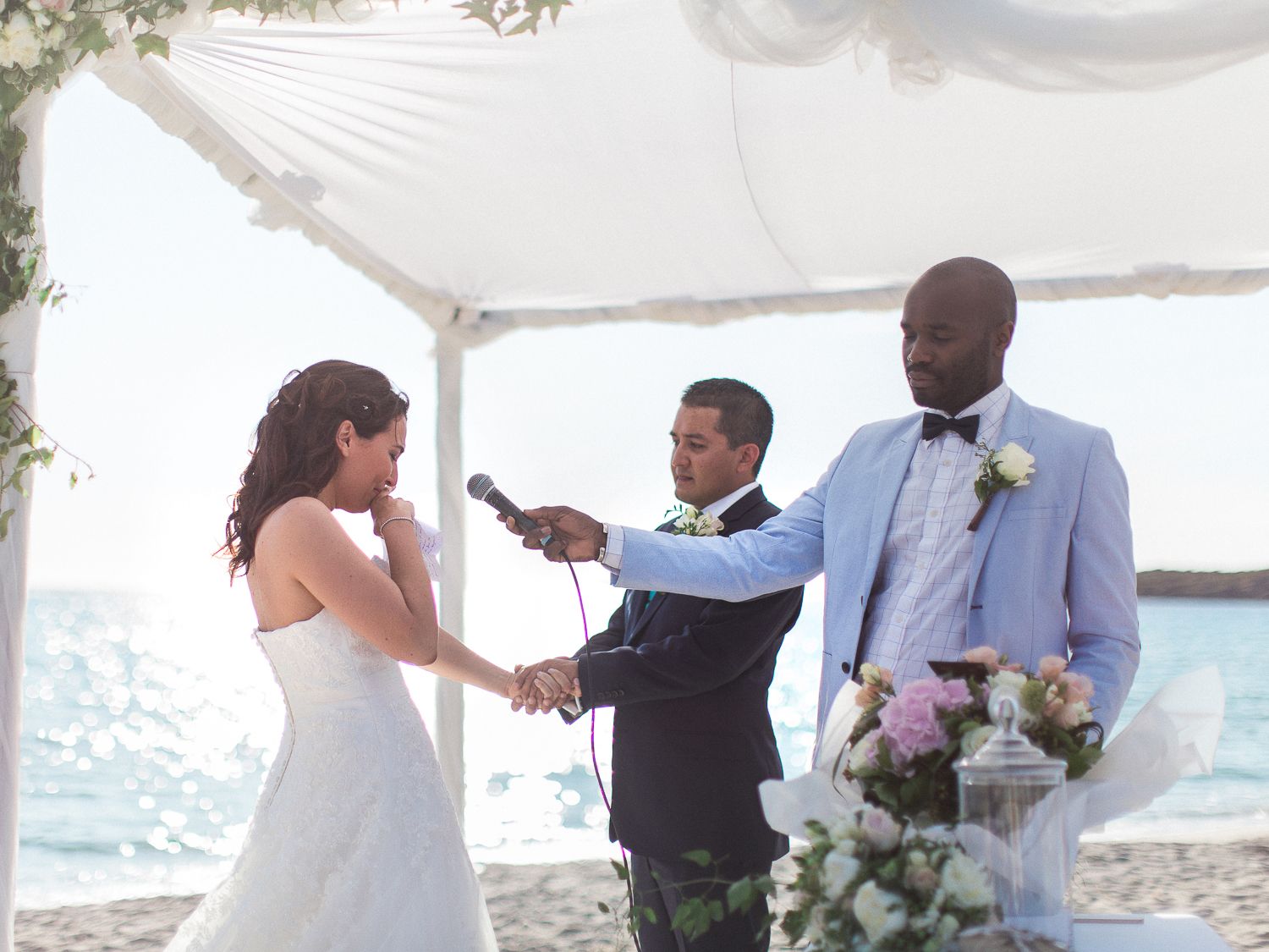 Un couple échange ses vœux de mariage lors d'une cérémonie sur la plage, sous un dais blanc, un officiant tenant un microphone.