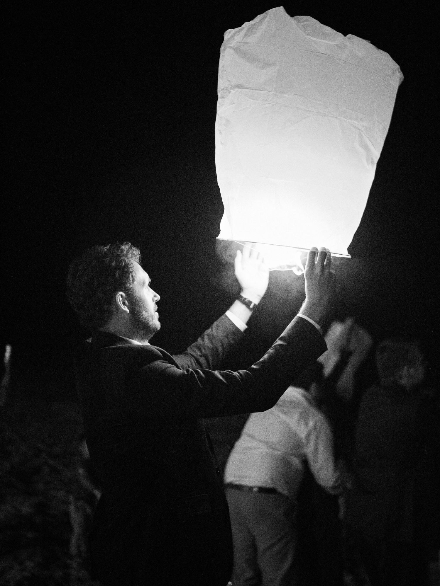 Photo en noir et blanc d'un homme en costume lâchant une lanterne céleste lumineuse dans le ciel nocturne à Ajaccio durant un mariage