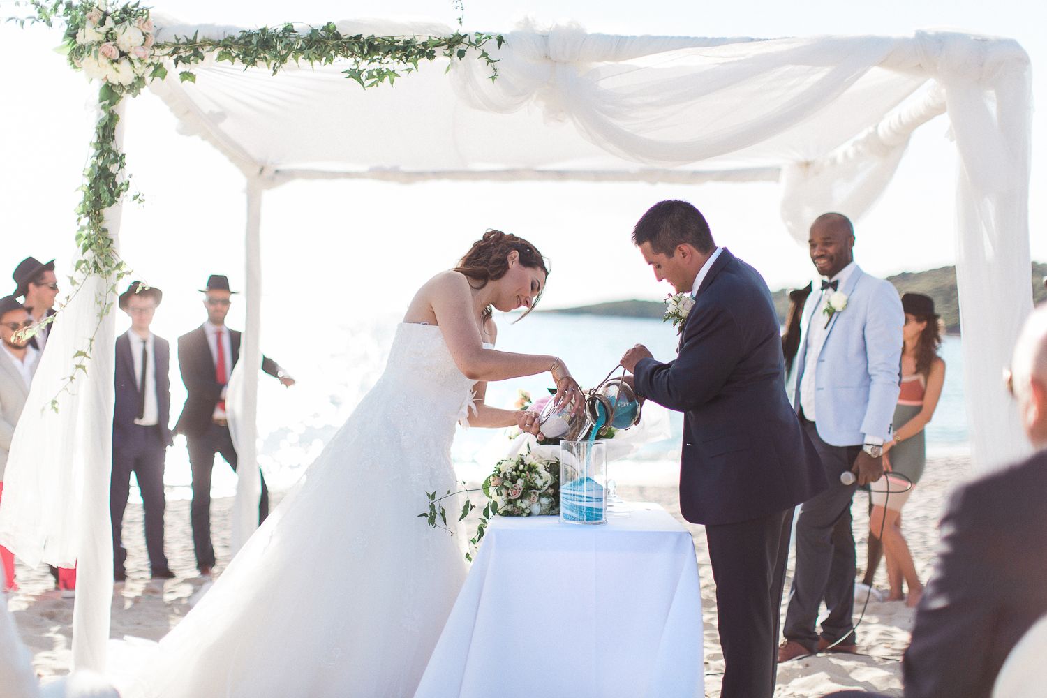 Les mariés échangent leurs alliances lors d'une cérémonie de mariage en plein air sur la plage, sous une canopée de fleurs blanches, sous le