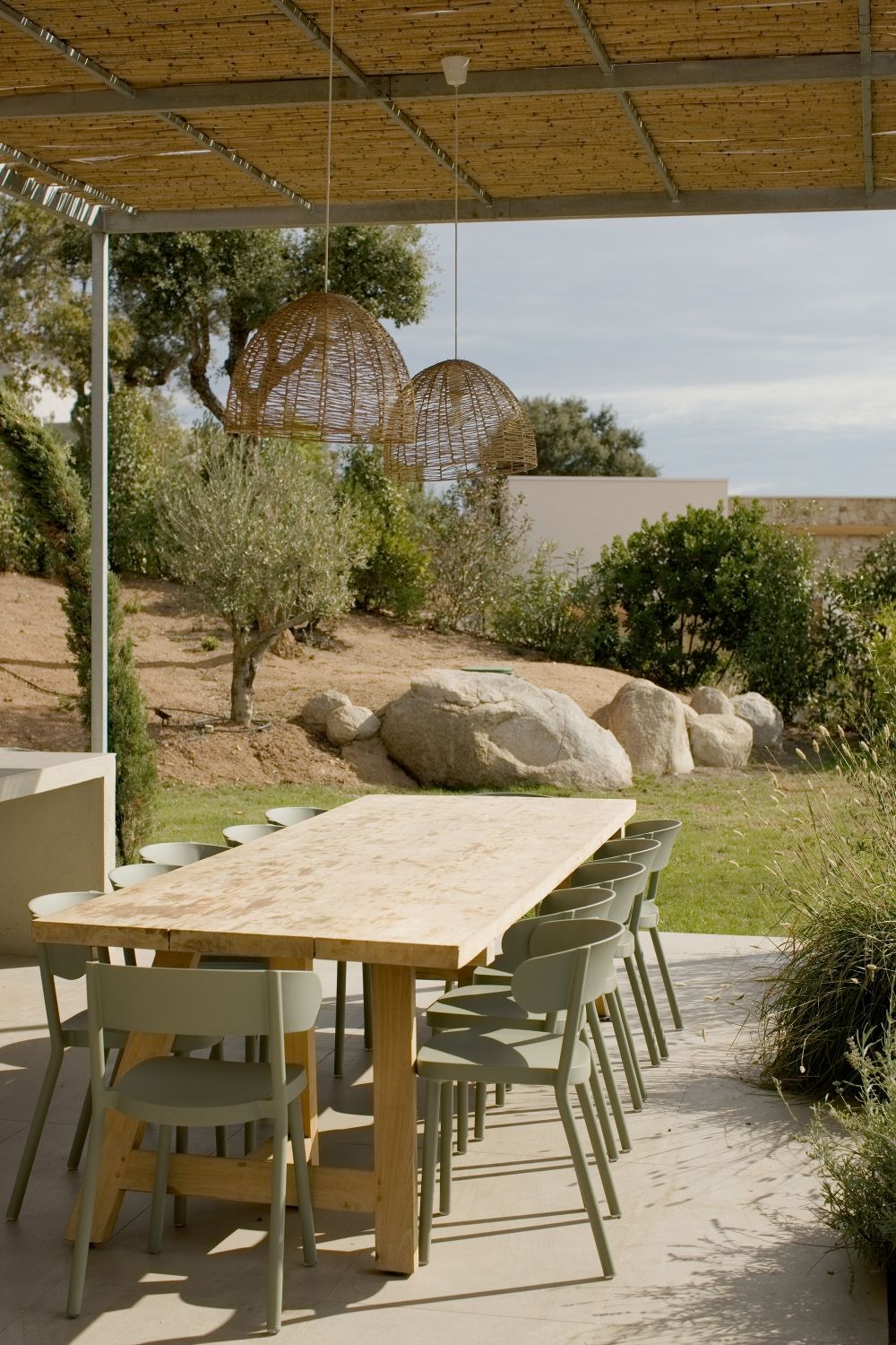 Espace repas extérieur rustique avec table en bois, chaises vert sauge, suspensions lumineuses en osier et aménagement paysager