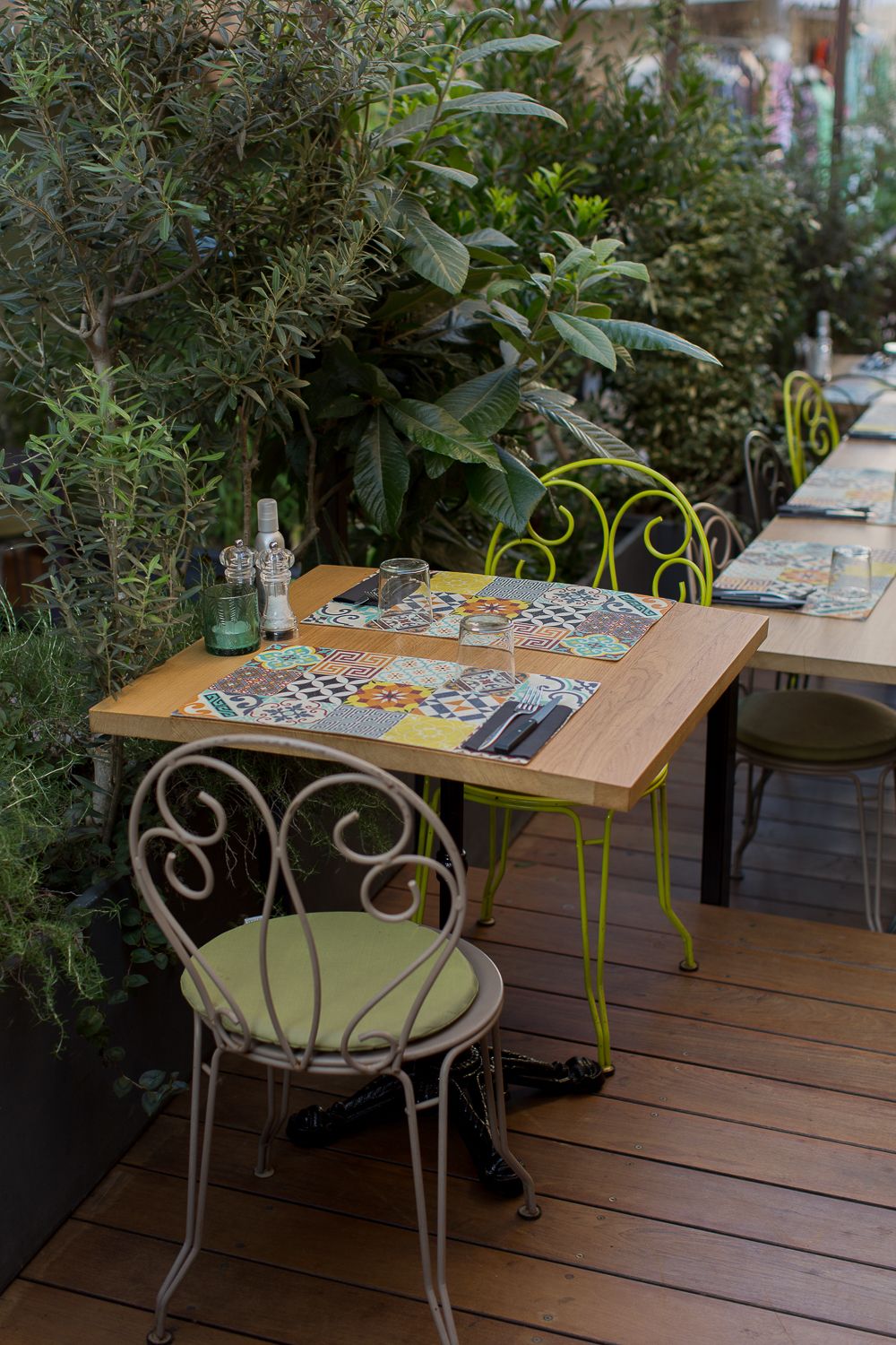 Terrasse extérieure de restaurant avec chaises en fer jaune, tables en bois, sets de table colorés et végétation luxuriante environnant