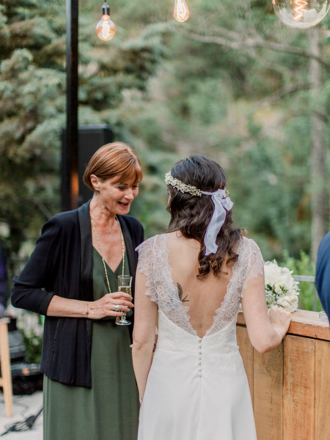 La mariée, vêtue d'une robe de mariée dos nu en dentelle et coiffée d'une couronne de fleurs, discute avec une invitée en robe verte