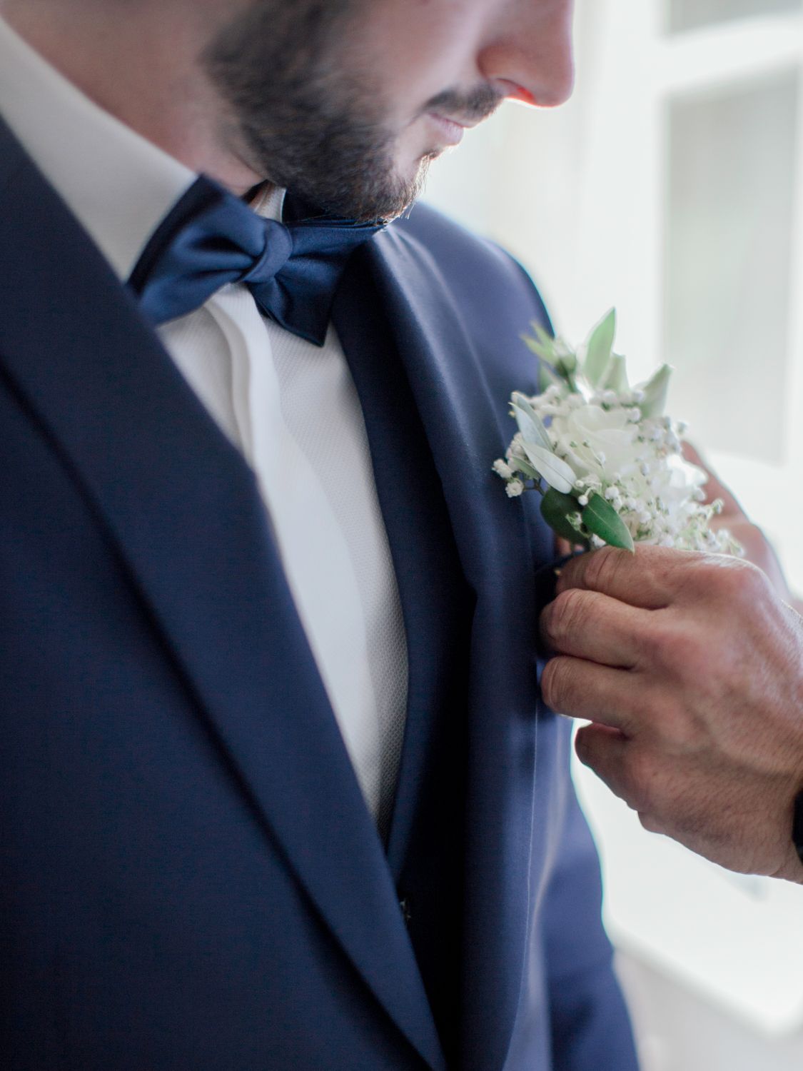 Close-up of groom in navy suit and bow tie having white floral boutonniere pinned on lapel.