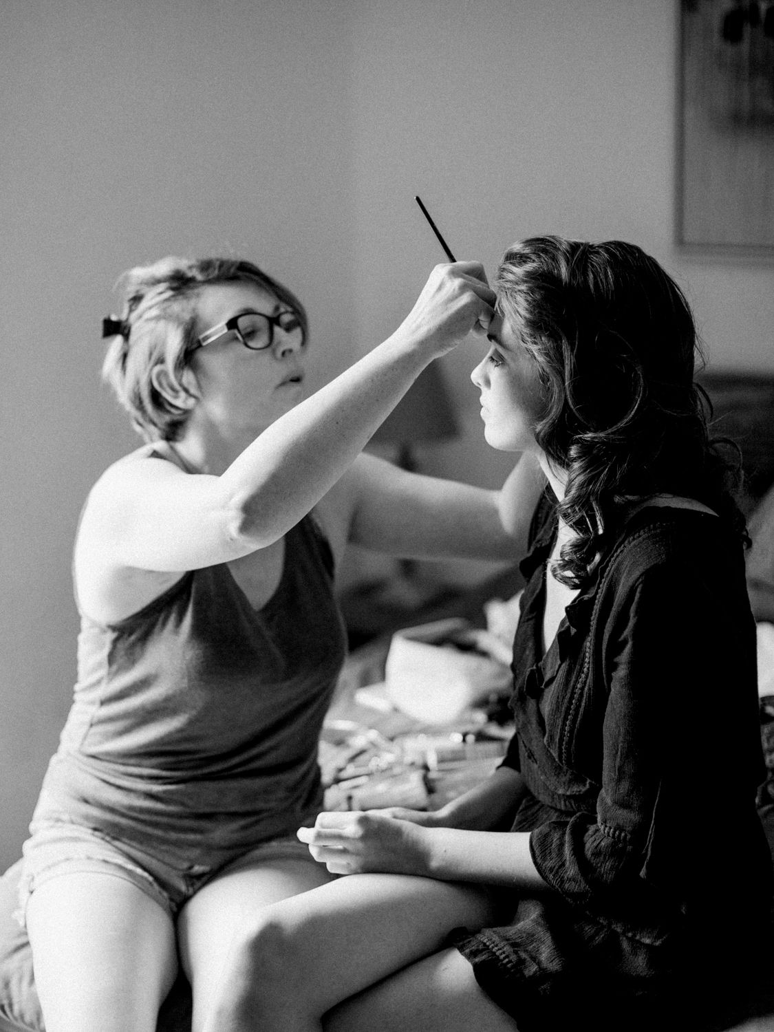 Black and white photo of a makeup artist applying eye makeup to a woman wearing glasses.
