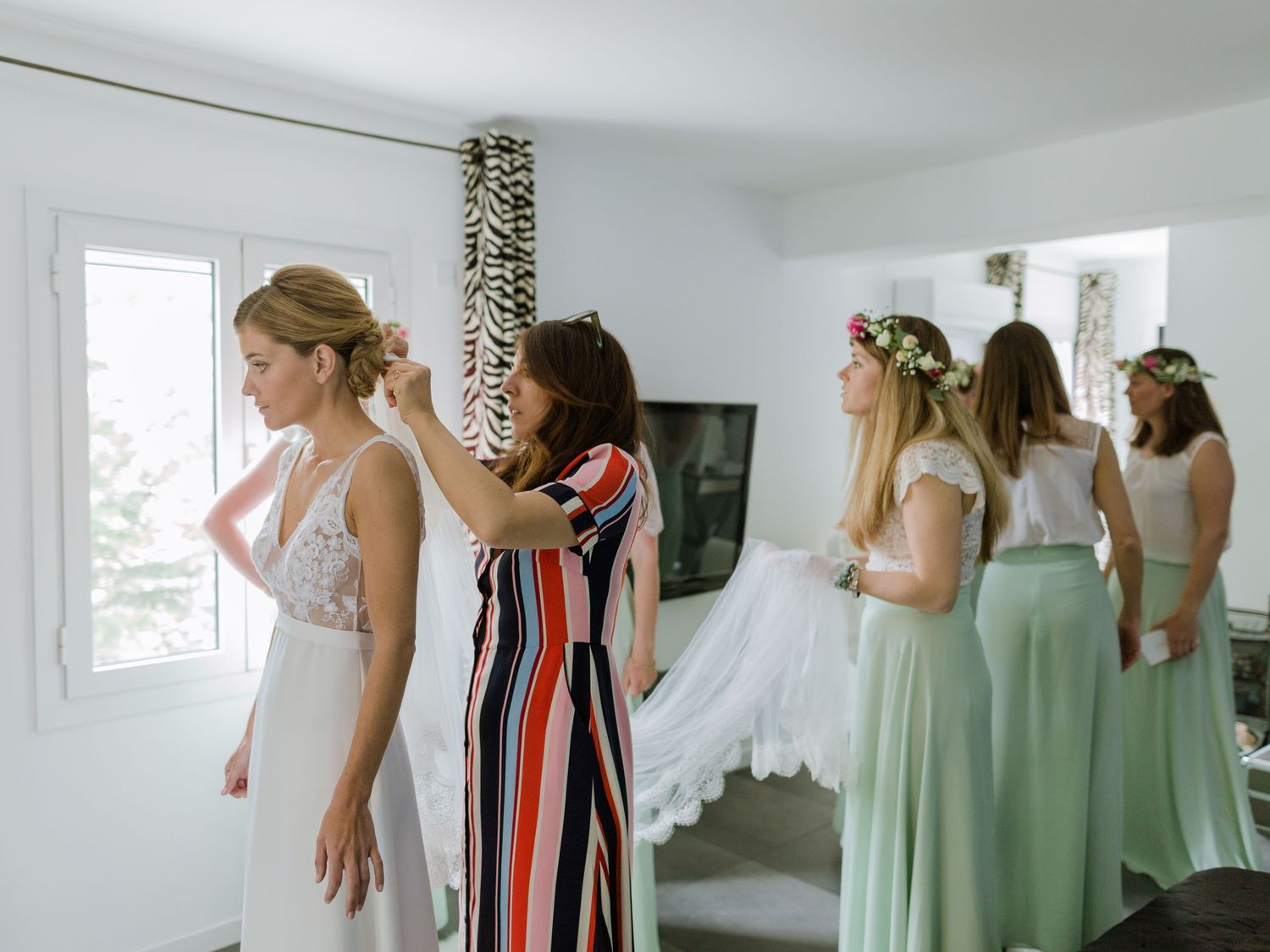 Dans une pièce d'un blanc éclatant, des demoiselles d'honneur en jupes vert sauge aident la mariée à se coiffer et à ajuster sa robe.
