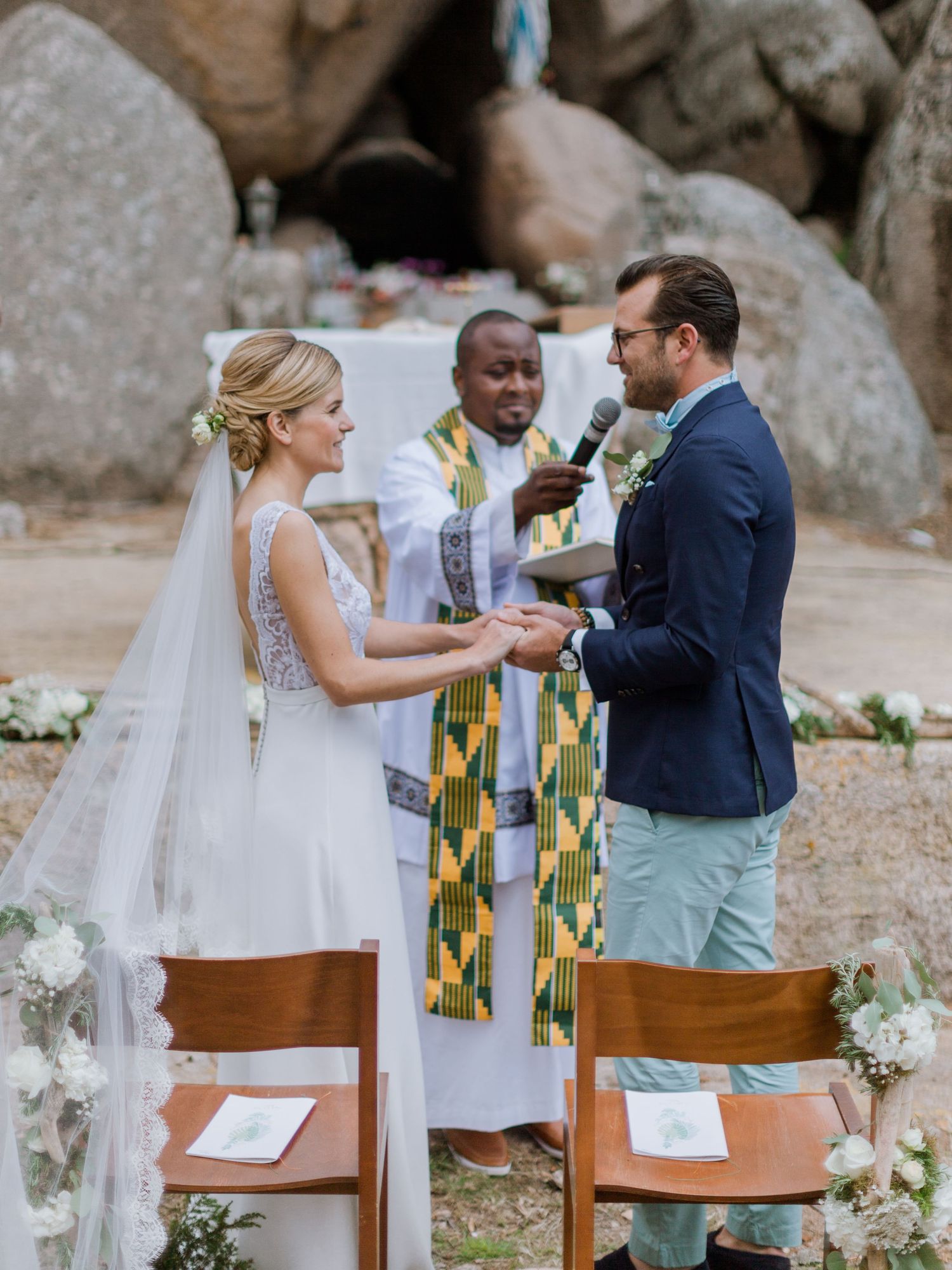 Les mariés échangent leurs vœux en plein air, l'officiant portant une étole colorée, devant un décor rocheux et des décorations florales.