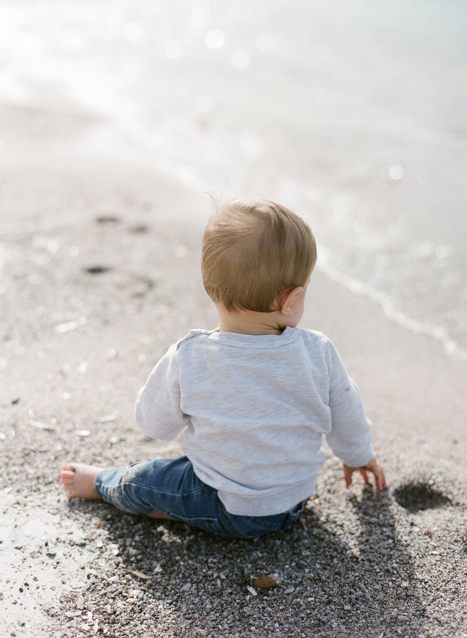 Un petit garçon en chemise grise et jean est assis sur une plage de sable fin face à la mer de Santa Giulia, porto-vecchio.
