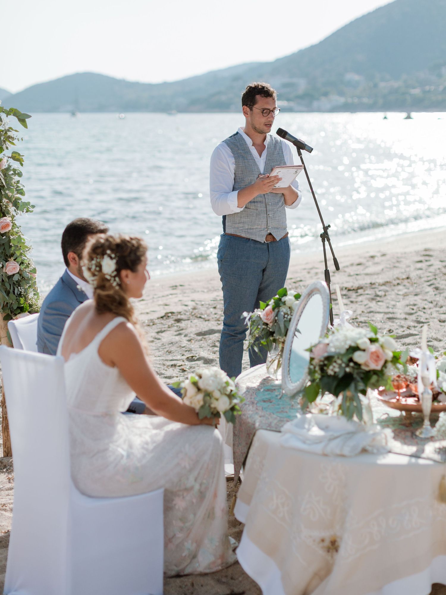 Un homme prononce un discours lors d'une cérémonie de mariage romantique sur la plage, avec les mariés assis au bord de l'eau.