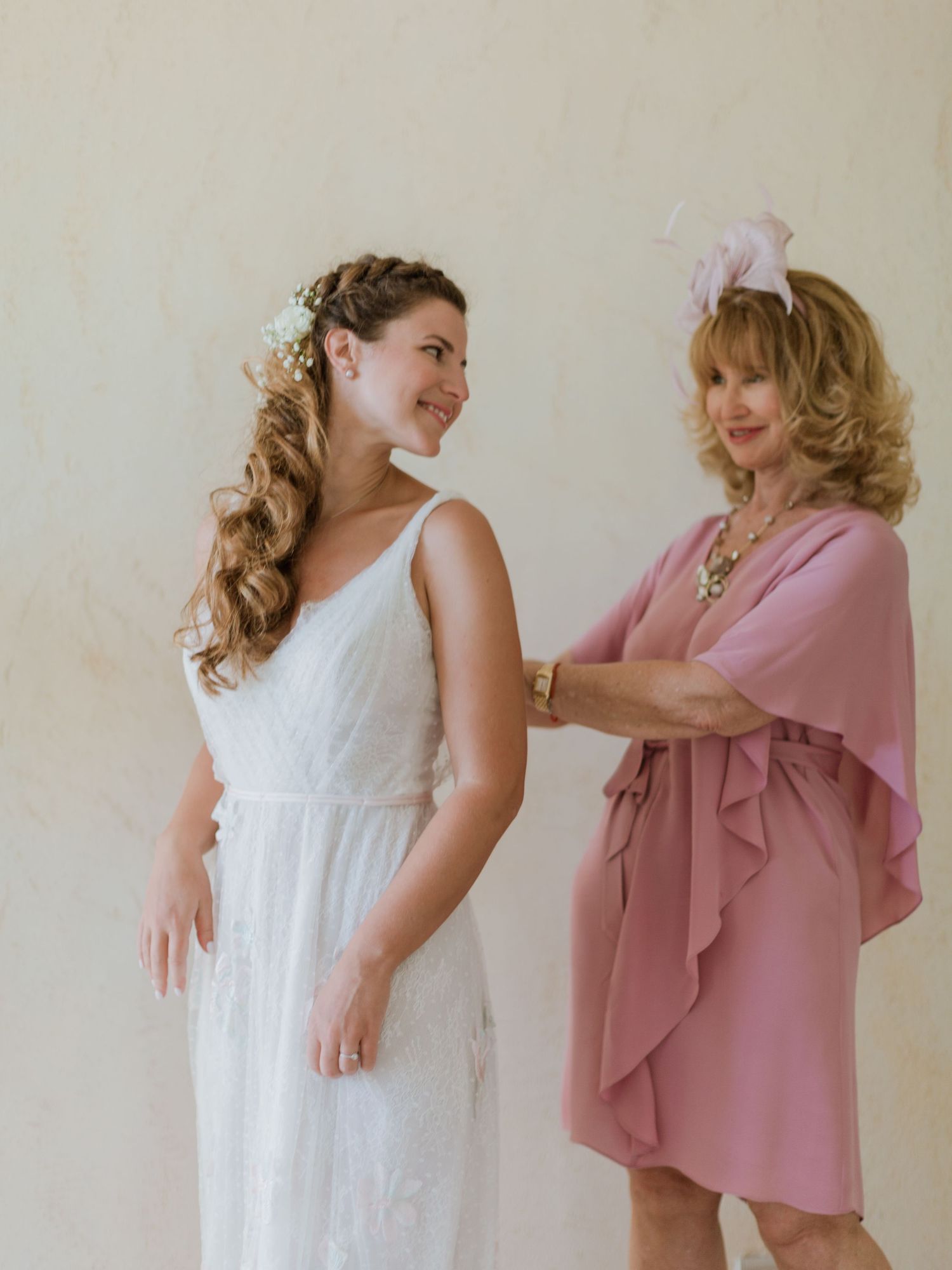 Une mariée en robe de dentelle blanche sourit tandis qu'une femme plus âgée en robe rose ajuste sa tenue avant la cérémonie de mariage.