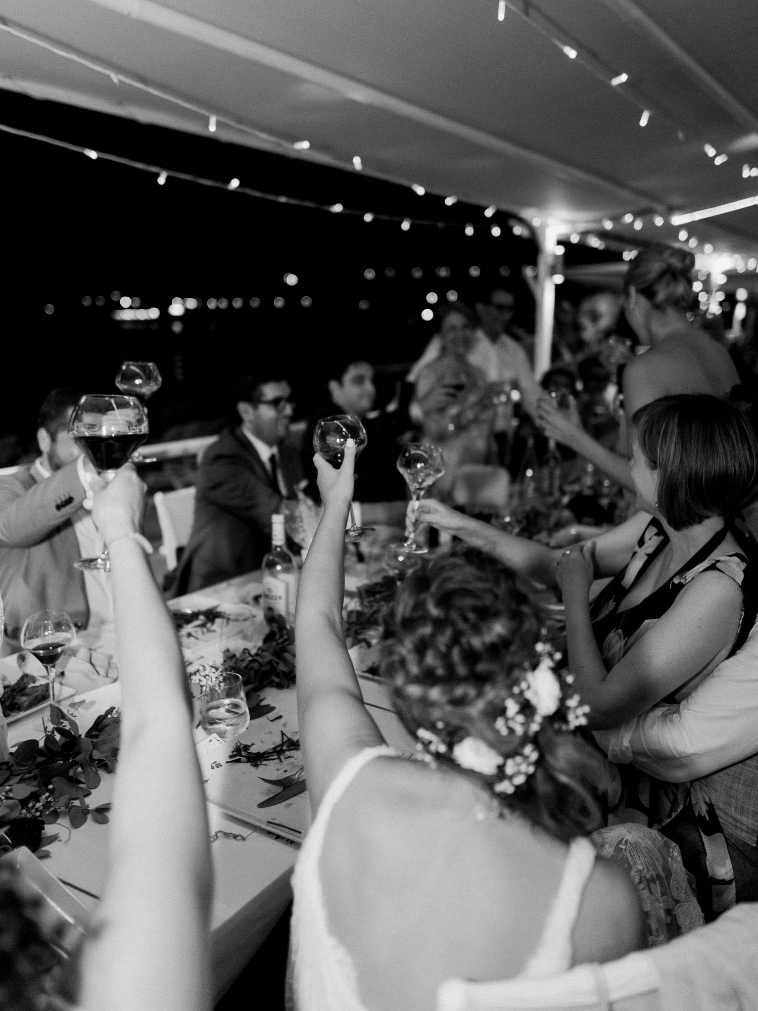 Photo en noir et blanc d'invités à un mariage levant leur verre pour un toast sous une tente décorée de guirlandes lumineuses.