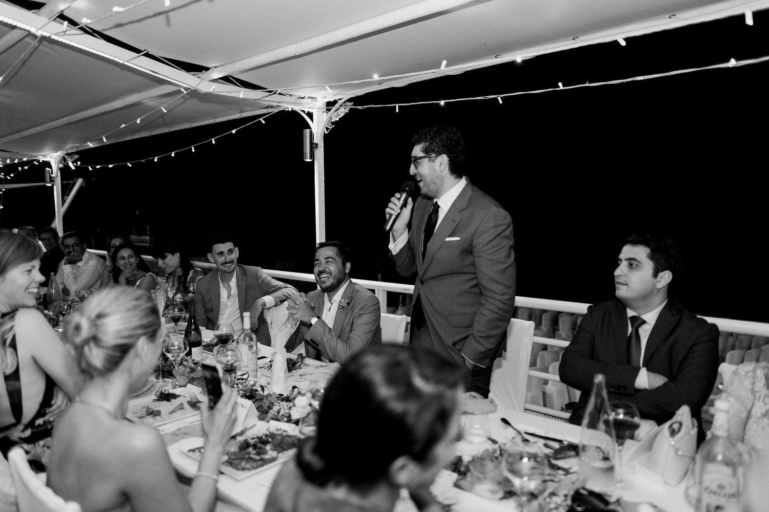 Un homme prononce un discours lors d'un élégant dîner de réception de mariage en plein air ; les invités sont assis à des tables décorées