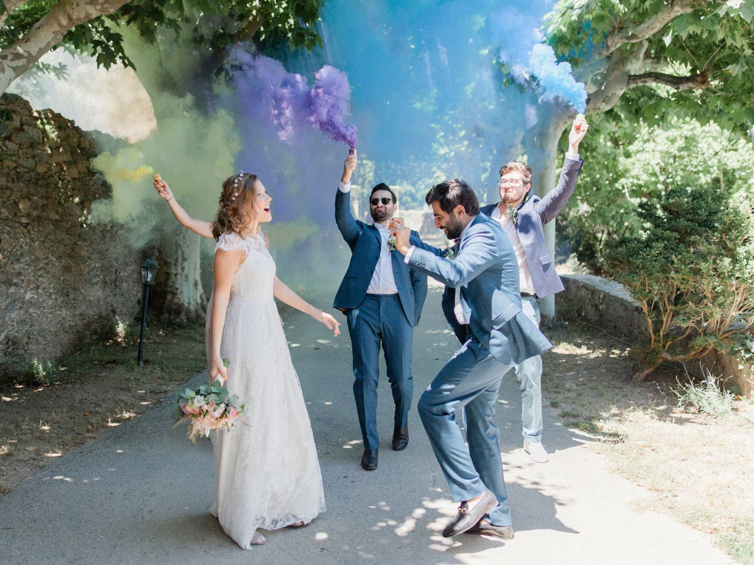 Joyful wedding party celebrating outdoors with colorful purple and blue smoke bombs on a sunny tree-lined path.