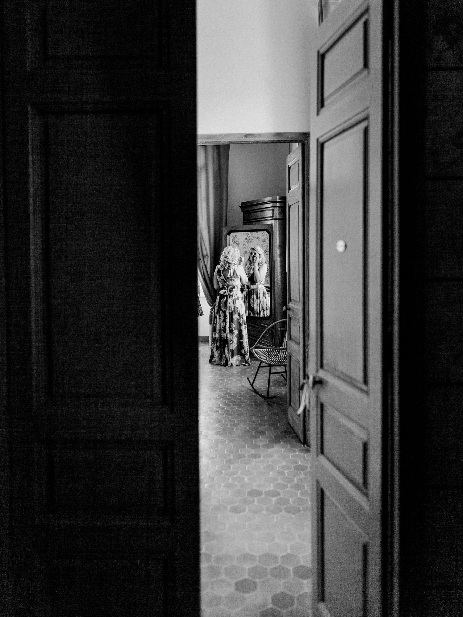 Black and white photo of a person reflected in a mirror through a doorway, with hexagonal tile floor and a chair nearby.