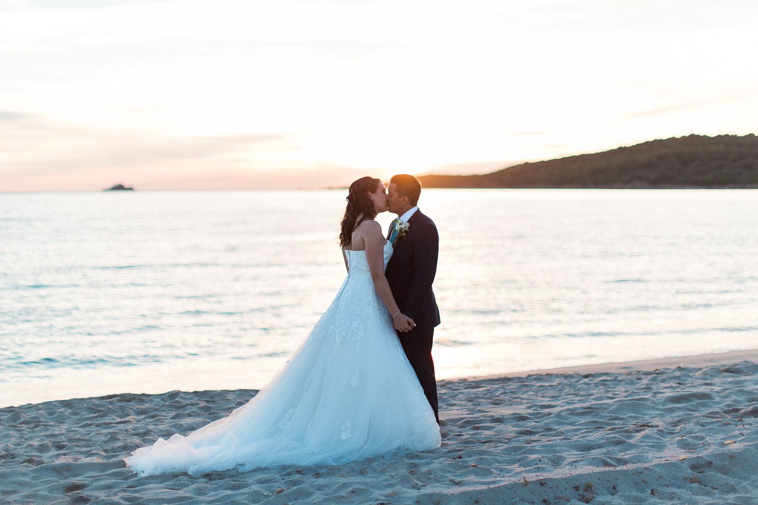Les mariés échangent un baiser romantique sur une plage de sable fin au coucher du soleil, avec les eaux calmes de l'océan en arrière-plan.