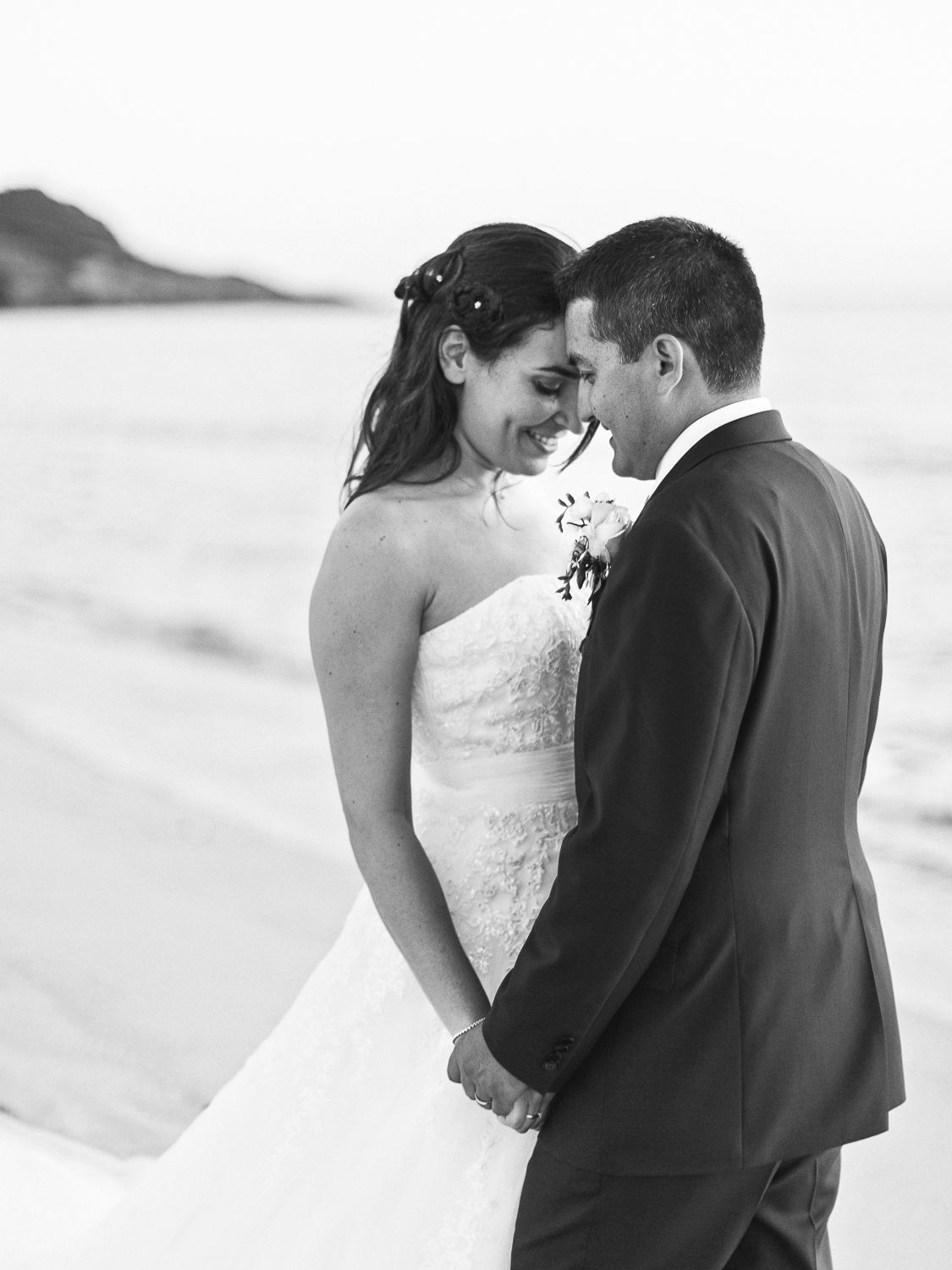Photo en noir et blanc d'une mariée et d'un marié se tenant la main et se touchant le front sur une plage.