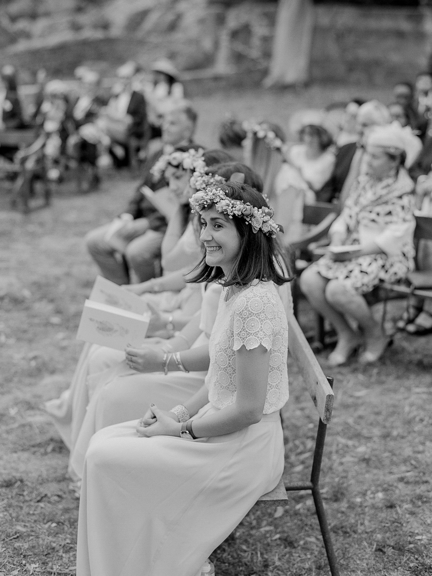 Photo en noir et blanc d'une demoiselle d'honneur souriante portant une couronne de fleurs, assise lors d'une cérémonie de mariage