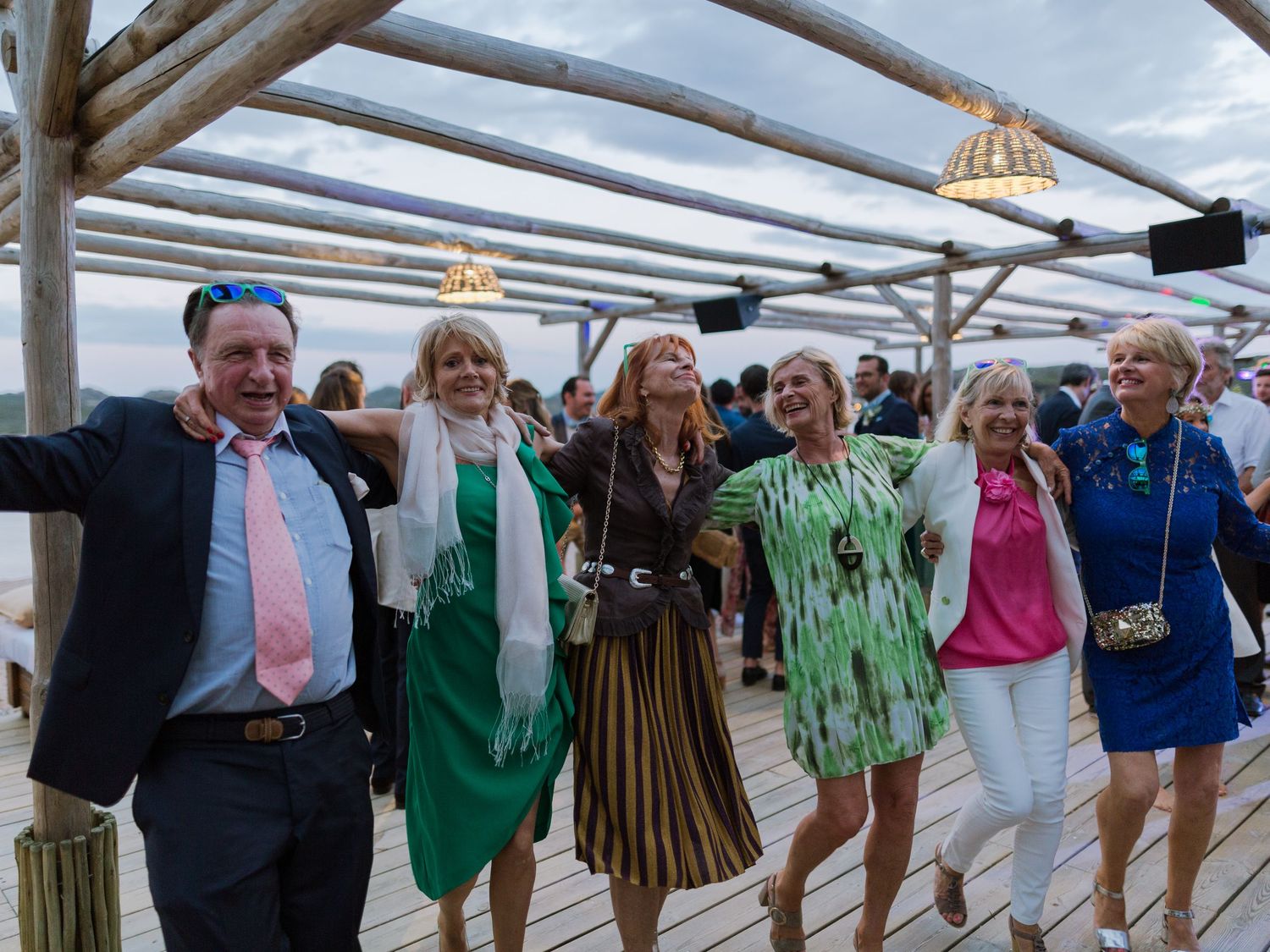 Un groupe de six adultes, vêtus de tenues de soirée colorées, célèbrent un événement festif en plein air sur une terrasse en bois.
