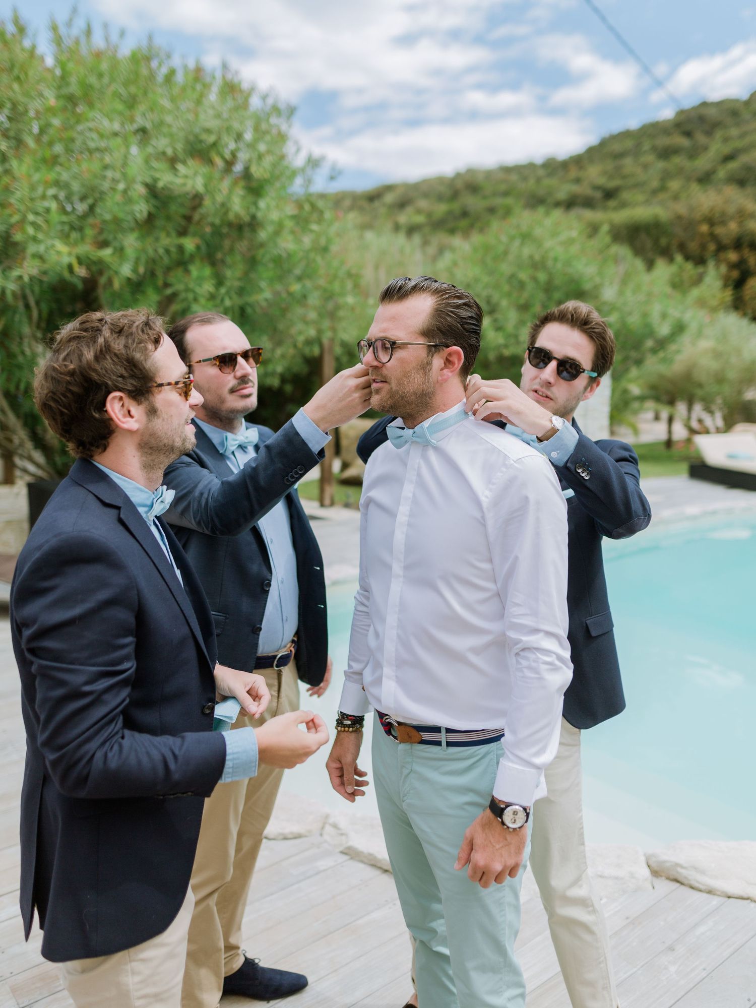Les garçons d'honneur en blazers bleu marine aident le marié à ajuster son nœud papillon et son col près de la piscine extérieure