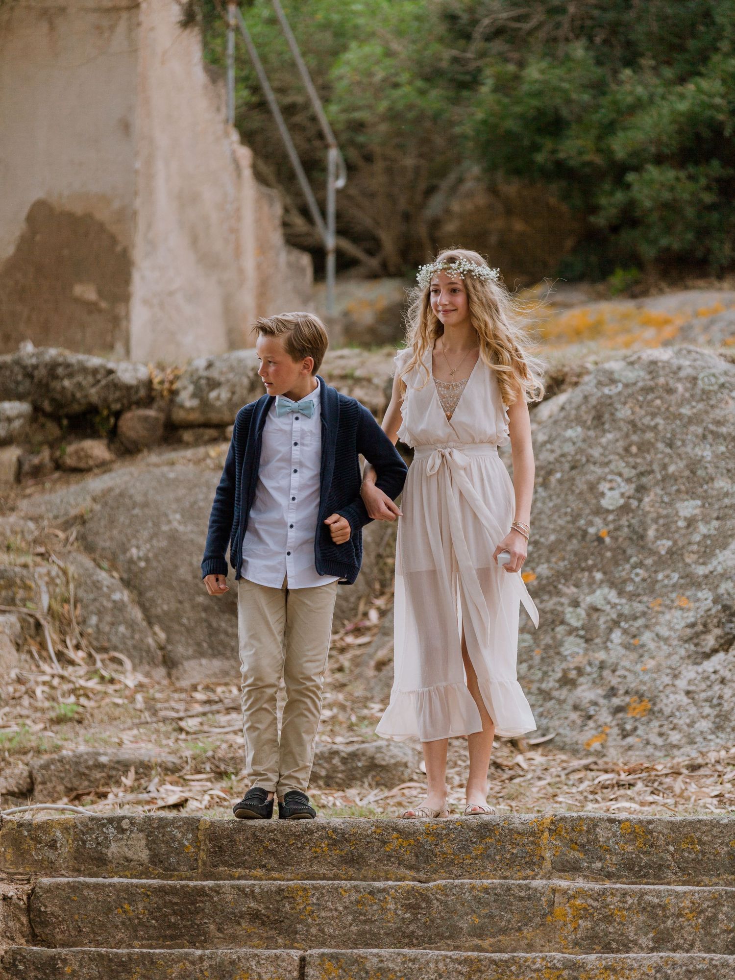Un garçon en blazer bleu marine et une fille en robe blanche fluide avec une couronne de fleurs se tiennent la main sur des marches
