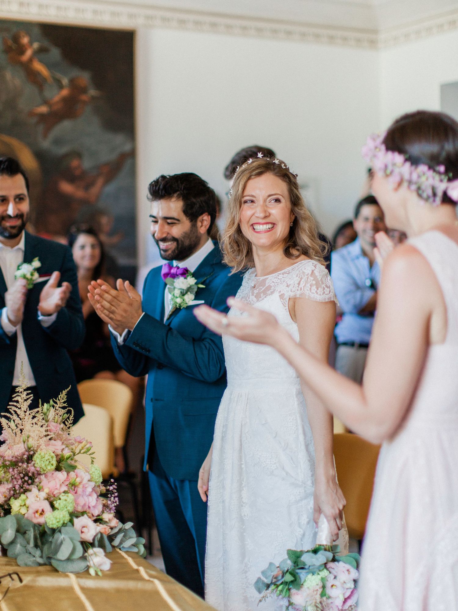 Bride and groom smiling and clapping during indoor wedding ceremony with floral arrangements and guests.