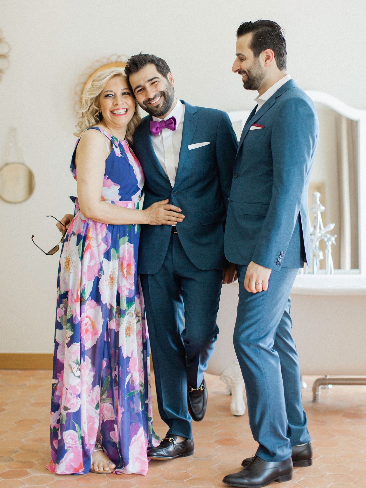 A woman in floral dress poses with two men in navy suits at a wedding celebration.