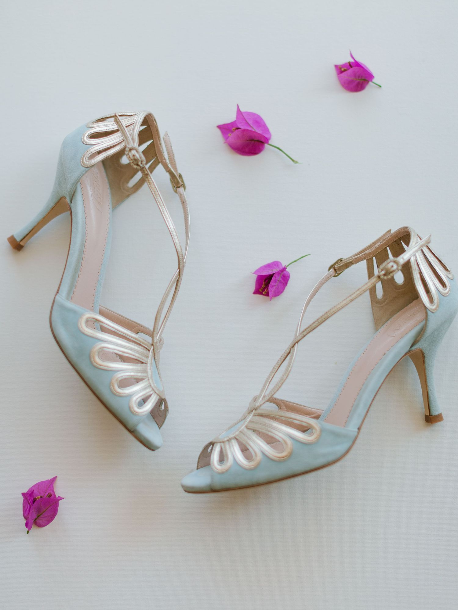 Sandales à talons et à brides, bleu clair et dorées, présentées sur une surface blanche parsemée de fleurs de bougainvillier roses.