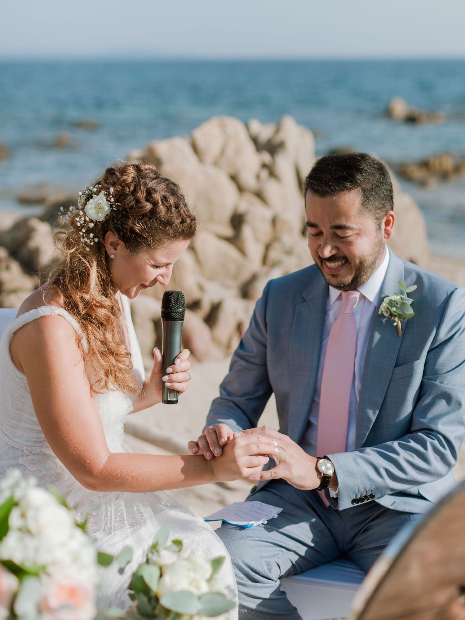 La mariée passe l'alliance au doigt du marié lors d'une cérémonie sur la plage ; tous deux sourient, avec l'océan et des rochers