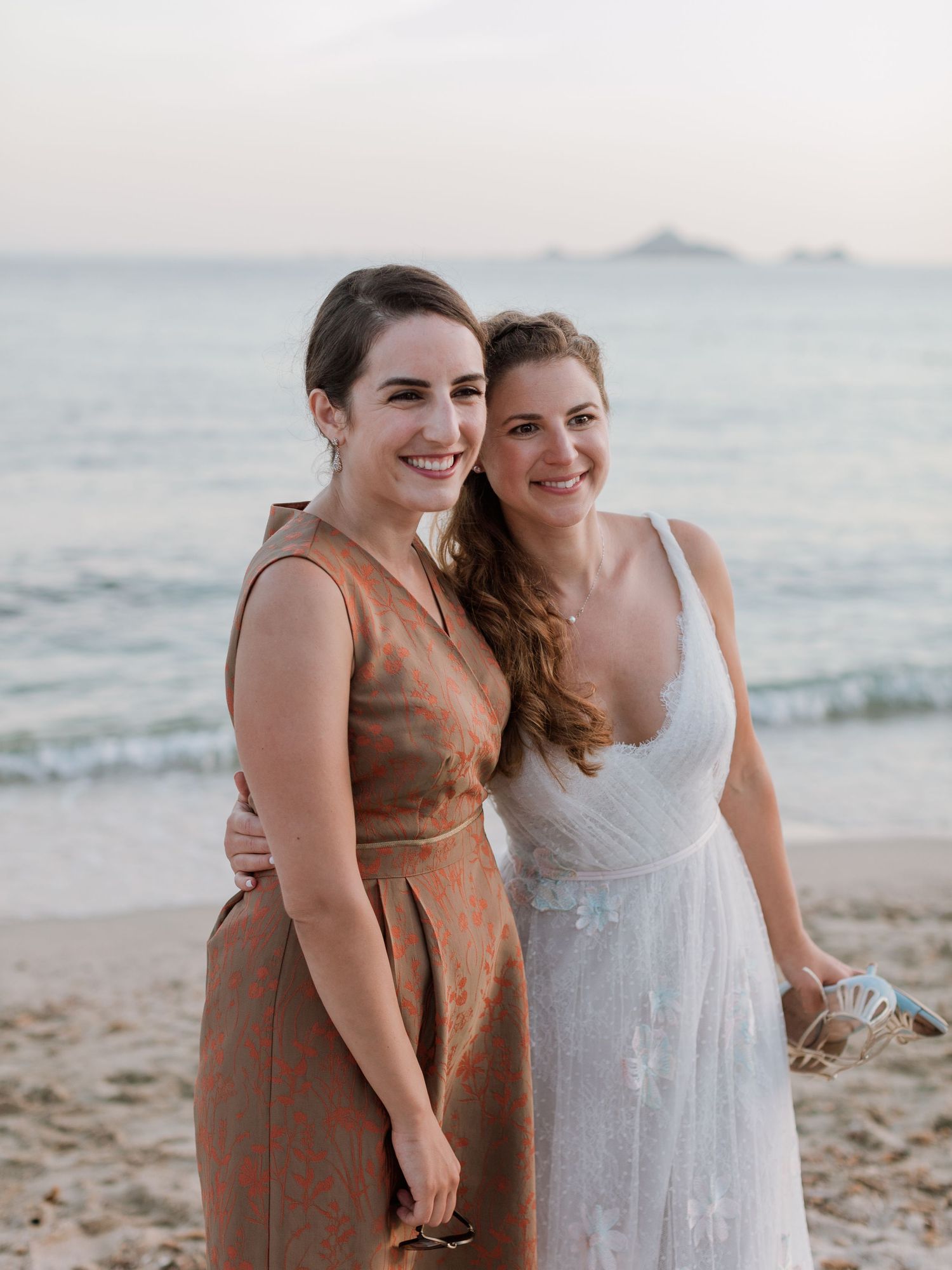 Deux femmes souriantes sur une plage au coucher du soleil, l'une en robe de mariée blanche et l'autre en robe couleur cuivre.