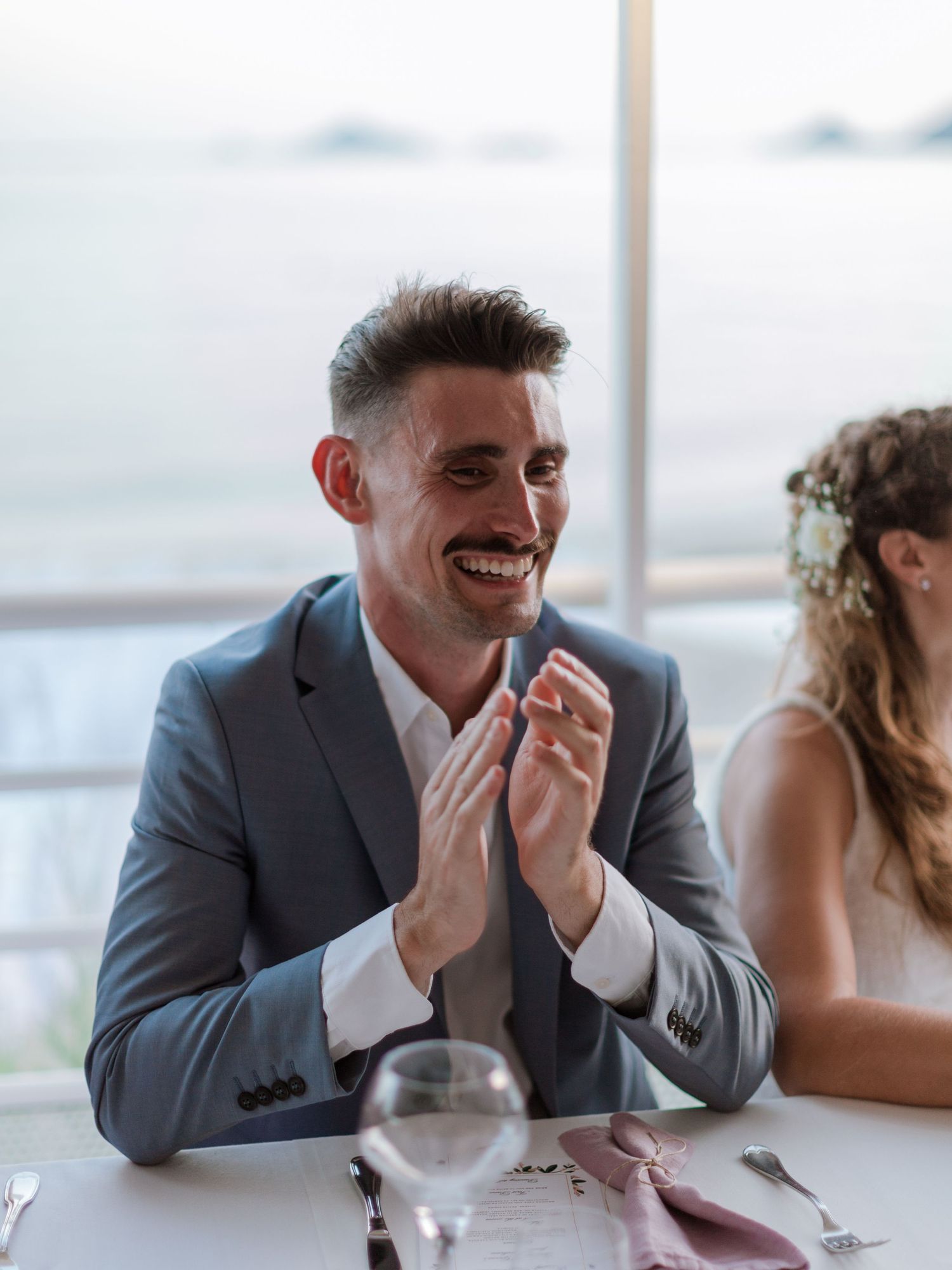 Marié souriant en costume gris applaudissant à une table de réception de mariage, avec la mariée à ses côtés et une vue sur l'océan