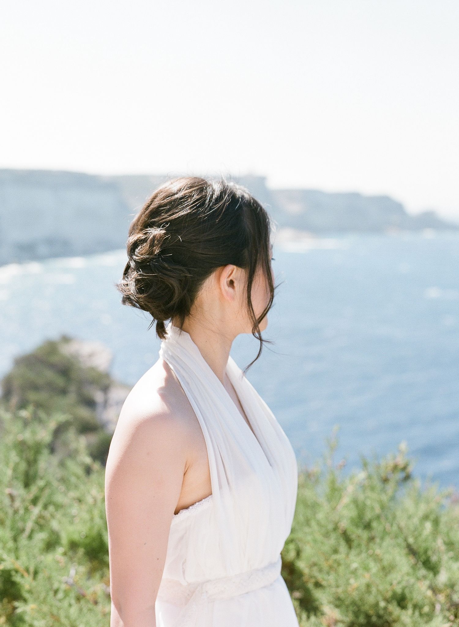 Une mariée, coiffée d'un chignon élégant et vêtue d'une robe blanche dos nu, contemple l'océan depuis le sommet d'une falaise pittoresque.
