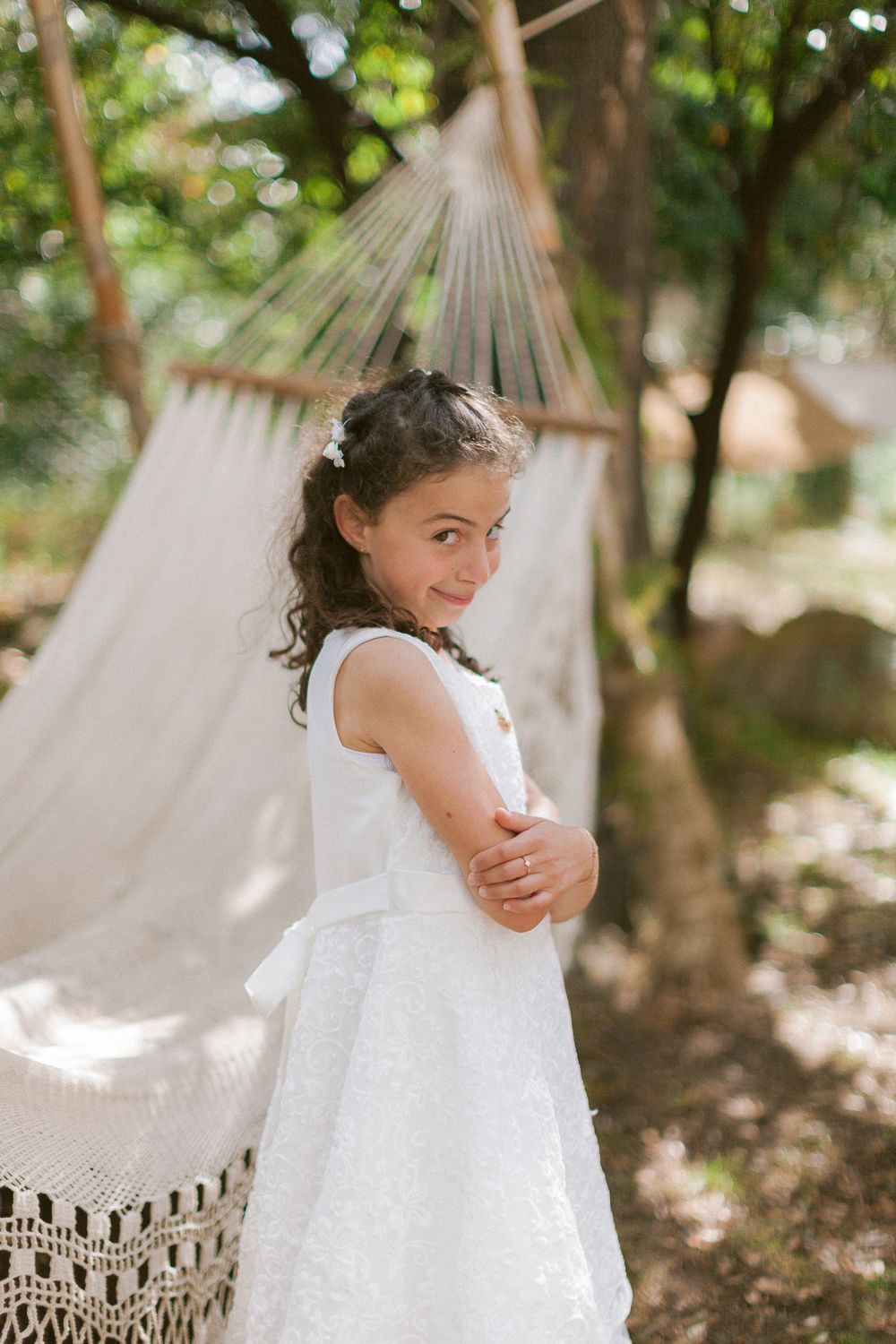 Jeune fille en robe blanche souriant par-dessus son épaule, avec un hamac et des arbres verts en arrière-plan, à Porto-Vecchio.