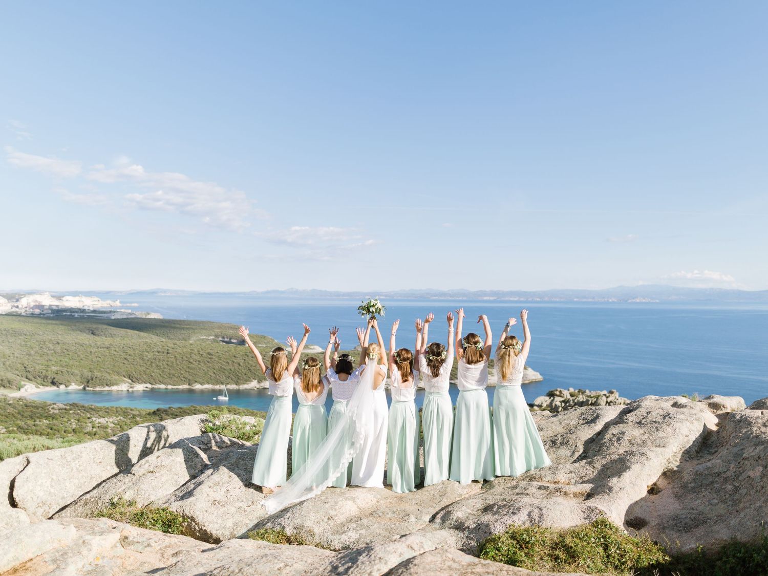 Bridesmaids in mint dresses raise arms on rocky cliff with stunning ocean and mountain backdrop.