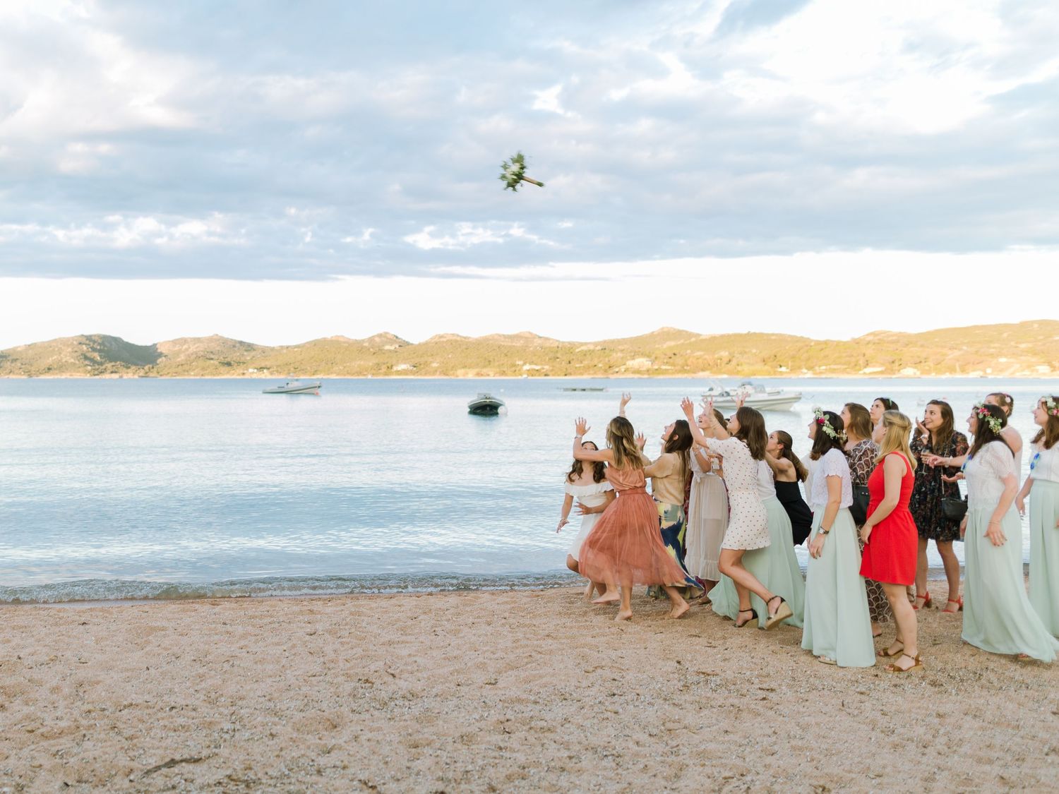 Des demoiselles d'honneur en robes colorées sont alignées sur une plage de sable fin tandis qu'un bouquet est lancé en l'air.