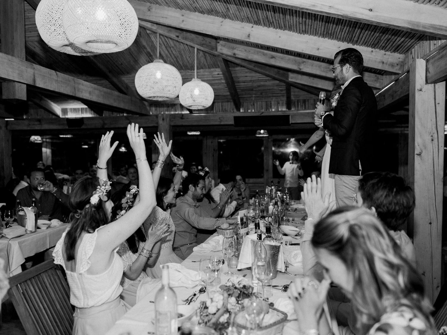 Photo en noir et blanc d'une réception de mariage montrant des invités levant la main tandis qu'un homme porte un toast