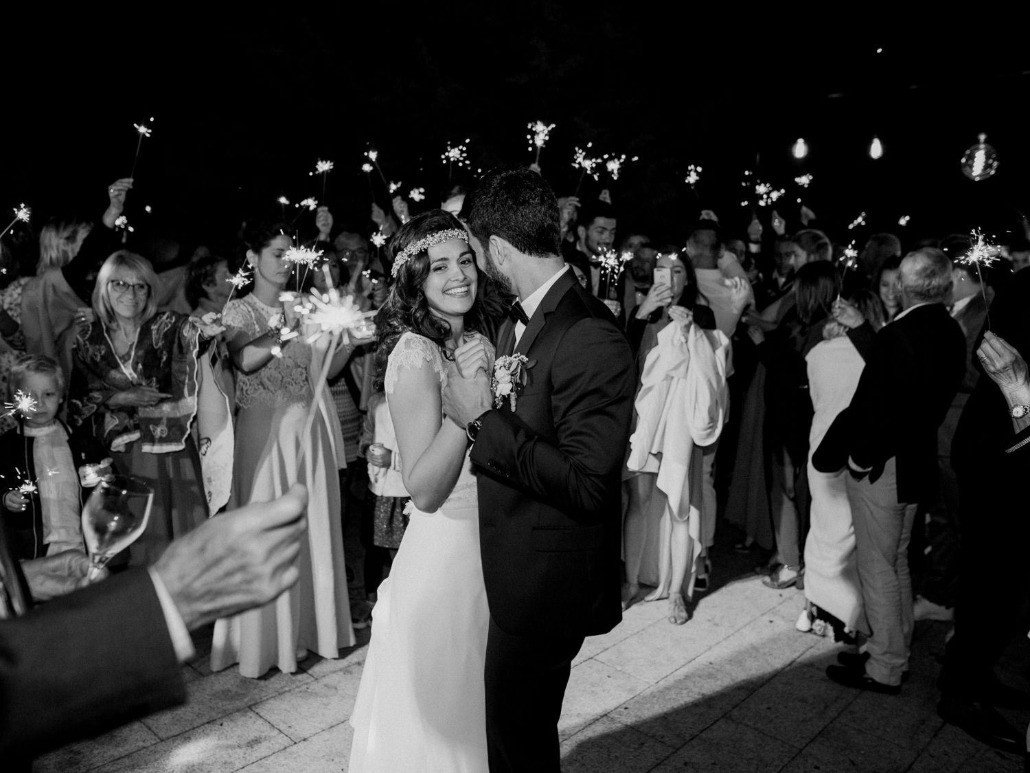 Photo de mariage en noir et blanc d'un couple s'embrassant, entouré d'invités tenant des cierges magiques, la nuit à Corte