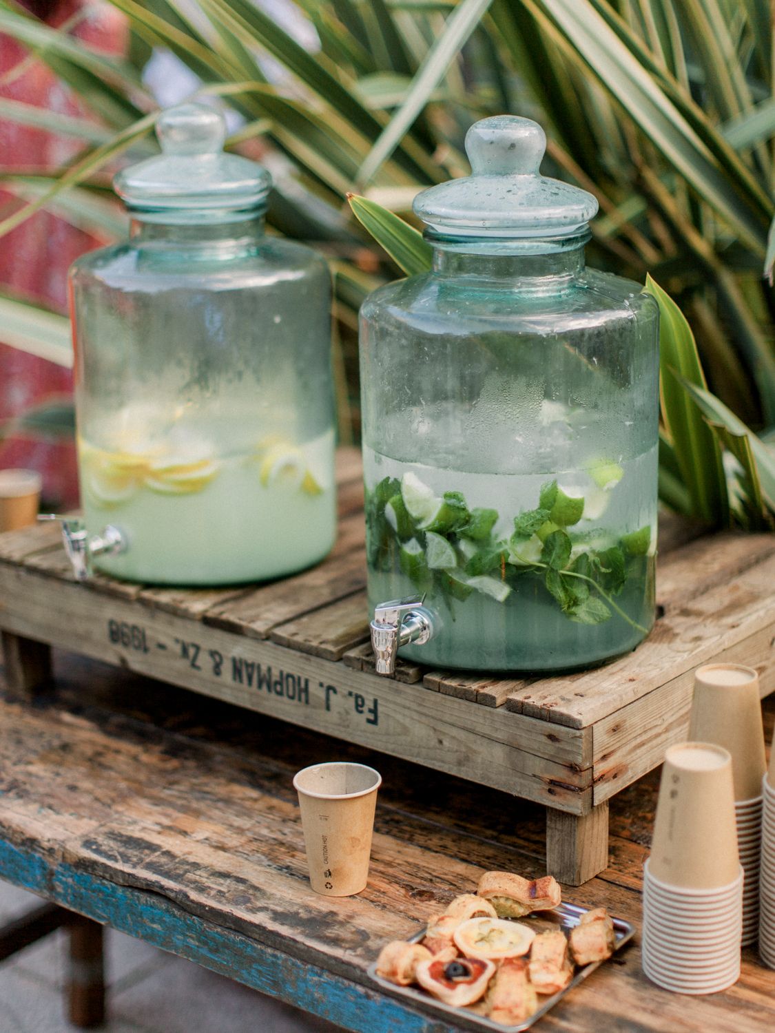 Deux fontaines à boissons en verre remplies d'eau infusée au citron et à la menthe, posées sur une table rustique en palettes de bois