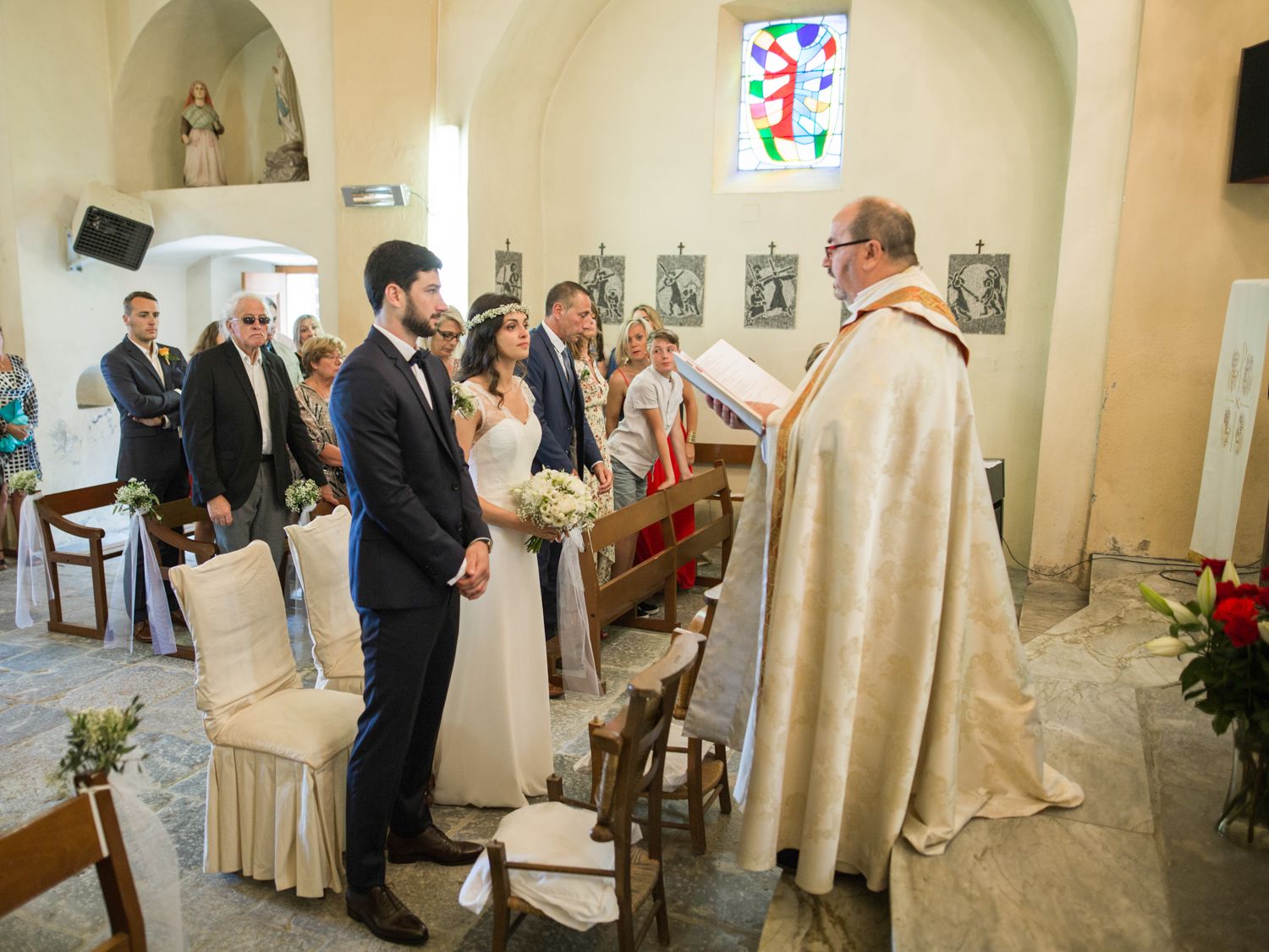 A couple exchanges wedding vows before a priest in a charming white church chapel with guests seated in rows behind them.